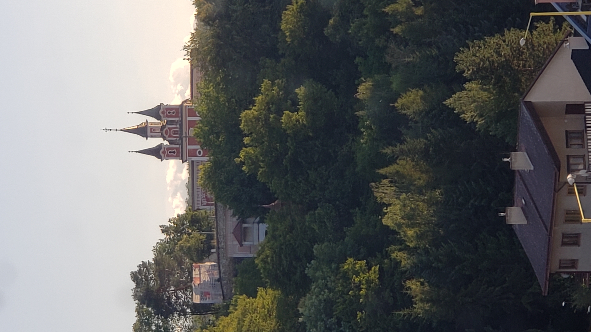 The image shows a panoramic view of the St. Nicholas Cathedral in Prešov, Slovakia, nestled atop a lush green hill. The cathedral, with its striking red and white facade and multiple spires, stands prominently against a clear sky. The foreground is dominated by a dense forest, with a glimpse of a residential building with a dark roof and chimneys in the lower right corner. The lighting suggests it is either early morning or late afternoon, casting a warm glow on the scene.
