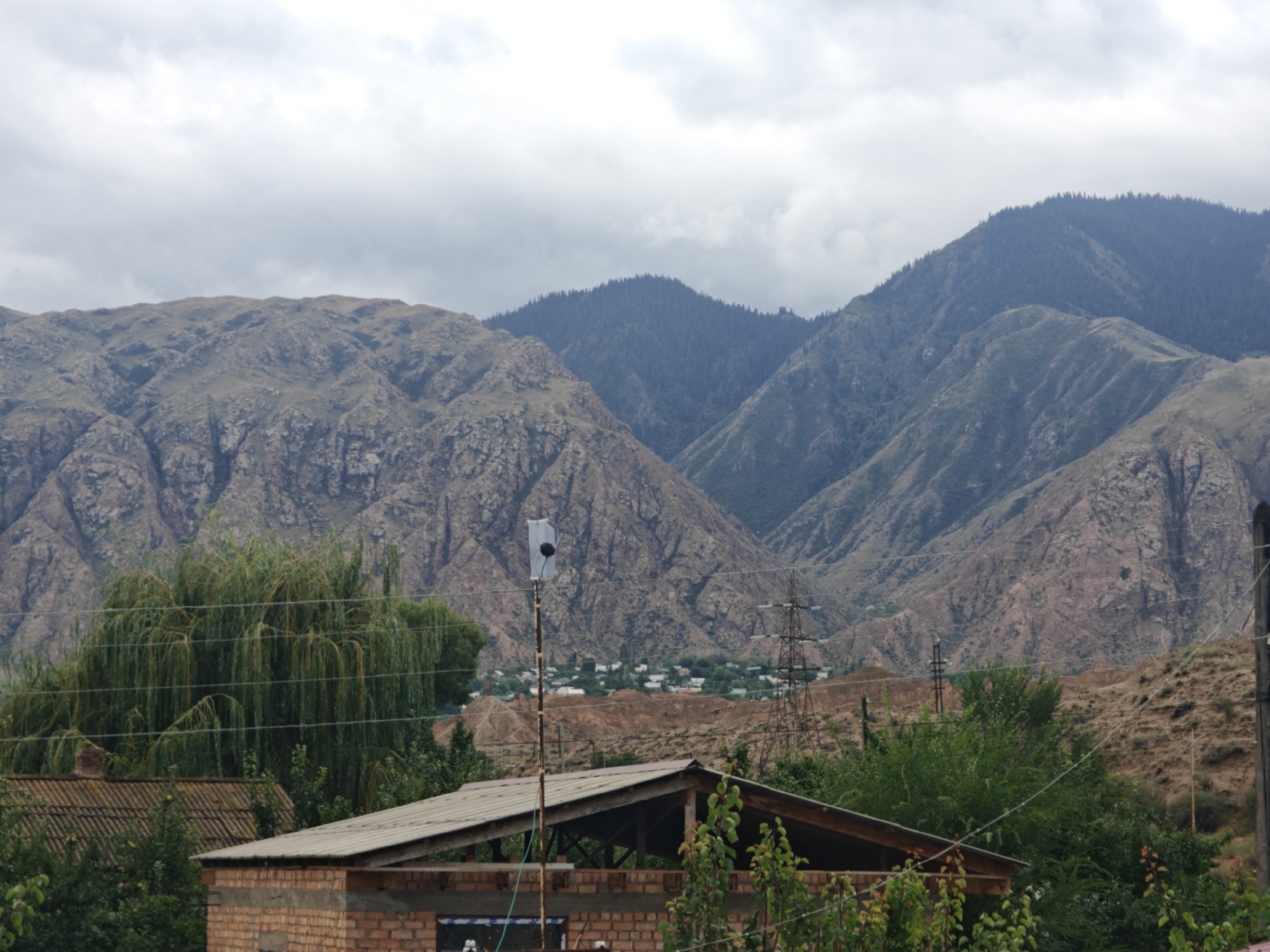 The image depicts a mountain landscape with a village in the distance, located in the vicinity of Каджи-Сай 2, Kyrgyzstan. In the foreground, there is a brick building under construction with a corrugated metal roof. A tall pole with a white object attached to the top stands to the left of the building. Several power lines traverse the scene. Beyond the building, a large, dense tree with hanging branches is visible, and further in the background, a small village is nestled among the hills. The background is dominated by rugged, layered mountains under a cloudy sky.
