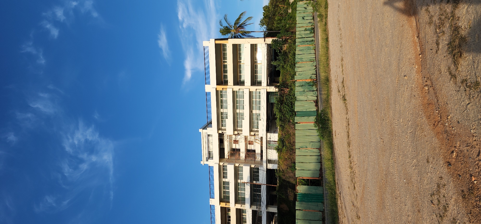 An exterior, ground-level, wide shot captures an unfinished, multi-story building in Ko Samui District, Thailand, under a clear blue sky with sparse white clouds. The building is modern in design, with several levels of balconies and white-paneled windows. Scaffolding is visible on one side, indicating ongoing construction or renovation. A palm tree stands to the right of the building. In the foreground, a temporary green metal fence runs horizontally, separating the building site from a dirt and gravel area. The ground appears dry and unpaved, with some patches of sparse grass. The overall impression is one of development or reconstruction in a tropical environment.