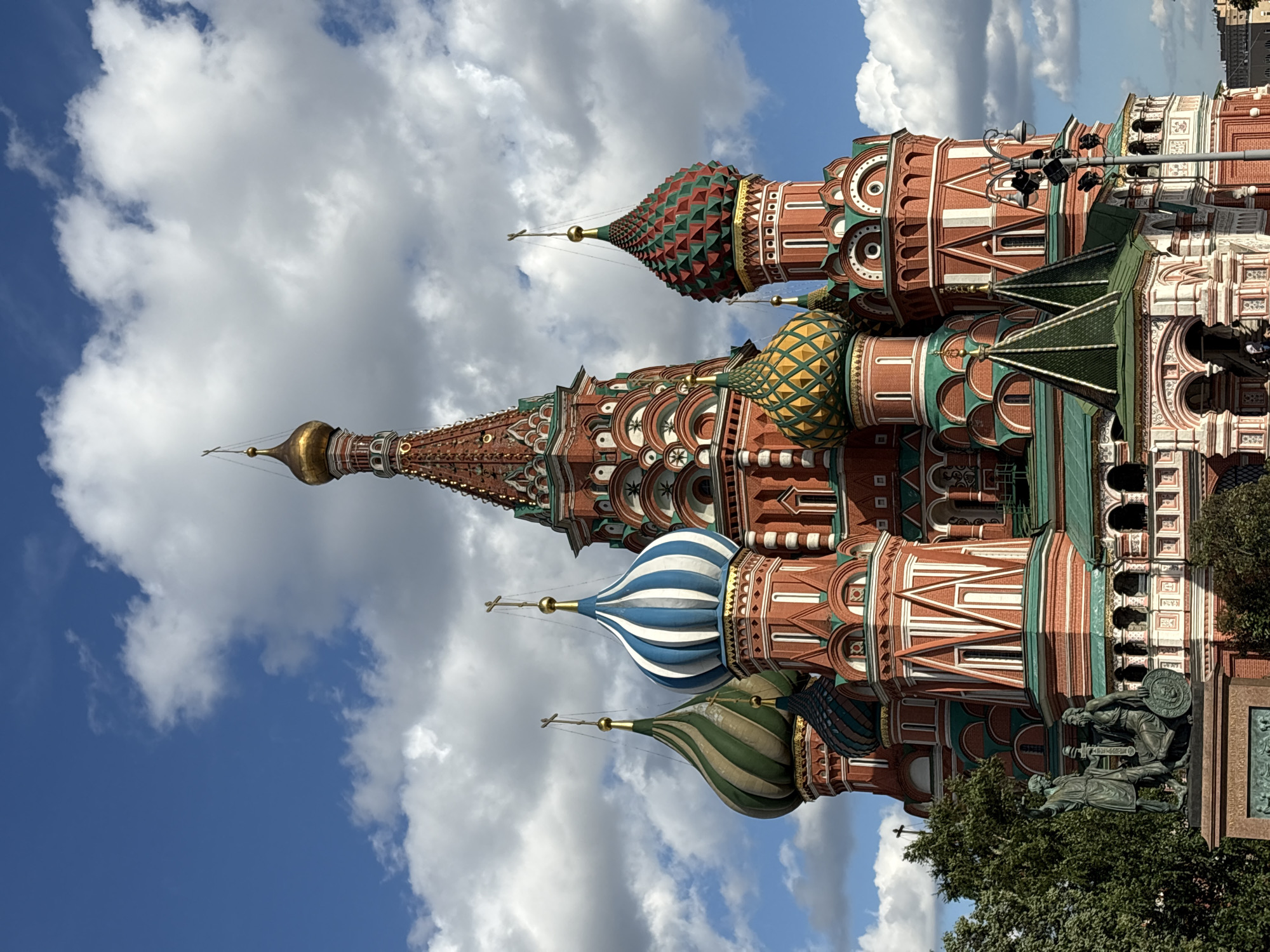 This is a close-up, eye-level, full shot of Saint Basil's Cathedral in Moscow, Russia, on a partly cloudy day. The iconic red brick cathedral with its distinctively colorful onion domes dominates the frame. The sky is a vibrant blue with scattered white, puffy clouds. Several domes are visible, including one with green and white stripes, another with green and yellow diamond patterns, and a central, taller dome with a golden spire. The cathedral's intricate architecture, characterized by red brick facades with white detailing and green roofs, is clearly visible. In the lower left corner of the image, a bronze statue of two figures, likely historical, is partially visible amongst lush green trees. The overall scene conveys a sense of grandeur and historical significance.
