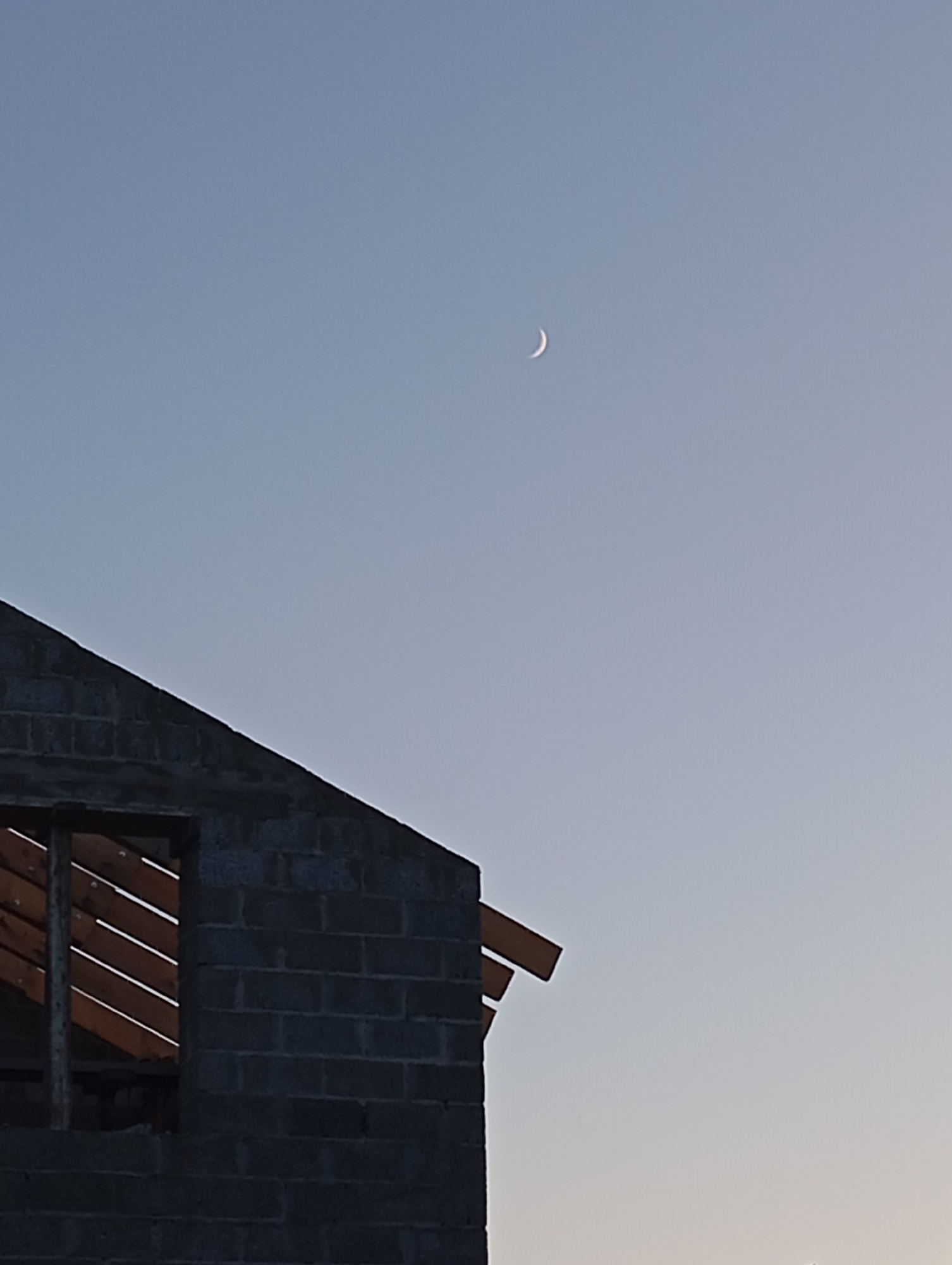 The image shows a crescent moon in a pale blue sky, with the unfinished roofline of a brick building in the foreground. The building appears to be under construction, with exposed wooden beams visible through a window opening. The sky is clear and there are no other objects or people visible in the frame. The overall impression is one of a serene, almost solitary scene, possibly taken at dawn or dusk. There is no information in the image to suggest the location is Orenburg, Russia.
