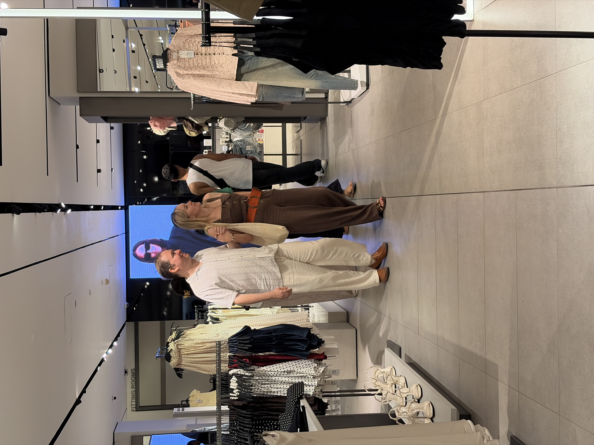 In a clothing store in Kuta, Indonesia, a man and a woman are walking together. The man is wearing a light-colored, short-sleeved collared shirt and loose-fitting cream pants. The woman is wearing a brown jumpsuit with an orange belt and holding a light-colored tote bag. They are walking past racks of clothing. In the background, a large screen displays an image of a person wearing sunglasses. The store appears to be well-lit with modern fixtures and displays.