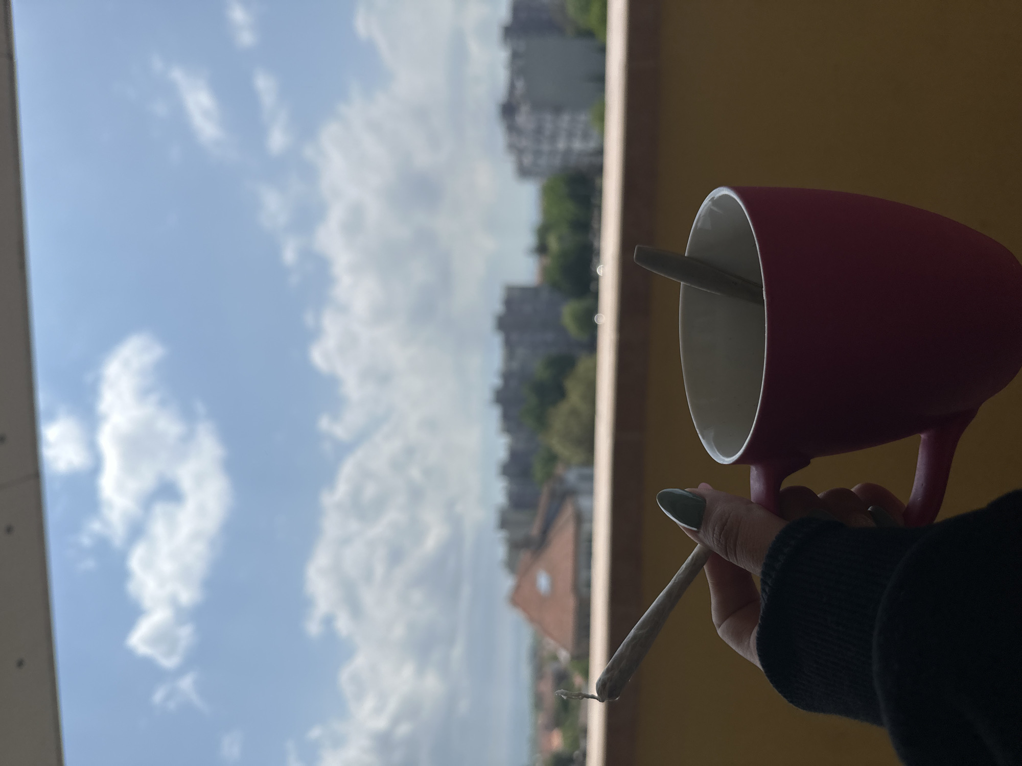 A person stands on a balcony, holding a lit hand-rolled cigarette in one hand and a maroon mug in the other, on a bright, sunny day in Burgas, Bulgaria. The hand holding the cigarette has a visible manicure with a dark green color. The mug appears to contain a spoon or similar utensil inside. The background features a cityscape with buildings and green foliage under a cloudy blue sky. The perspective suggests the photograph was taken at an elevated level, providing a view of the surrounding area.