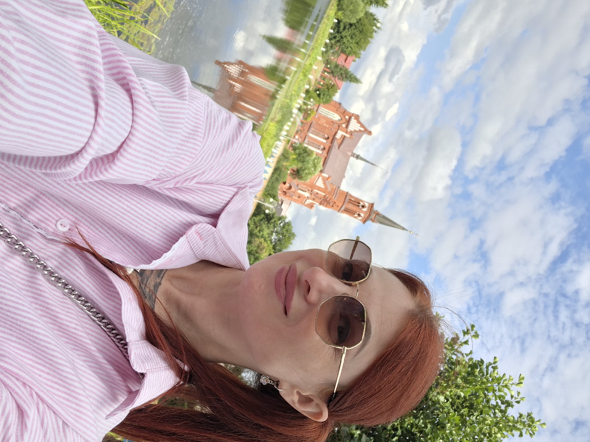 The image shows a woman with red hair, wearing sunglasses and a pink and white striped button-down shirt. She is taking a selfie outdoors, with a body of water and a red brick church in the background. The church appears to be Gothic in style, with spires and arched windows. The water reflects the church and the sky, which is partly cloudy. Trees and greenery surround the church and the water. This scene is likely located in the city of Pastavy, Belarus, given the architectural style of the church and the surrounding landscape.