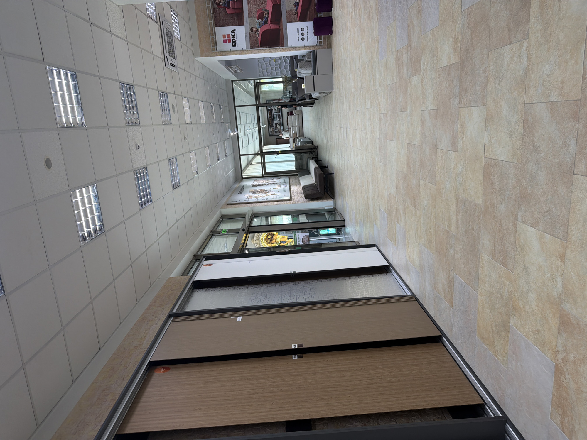 This is a wide, eye-level, interior shot of a furniture store in Trușeni, Moldova. The store features a long hallway with light-colored tile flooring. On the left, there are several large, display doors, some with wood grain patterns and others with decorative glass. Further down the hallway, more furniture is displayed, including sofas and chairs. The ceiling is a suspended grid with fluorescent lighting. Through a large glass partition at the end of the hall, more of the store's interior is visible, including additional furniture displays and a few people walking around. A large advertisement banner with "EDKA" prominently displayed is visible on the right wall, along with other promotional images. The scene appears to be a well-lit, organized retail environment.