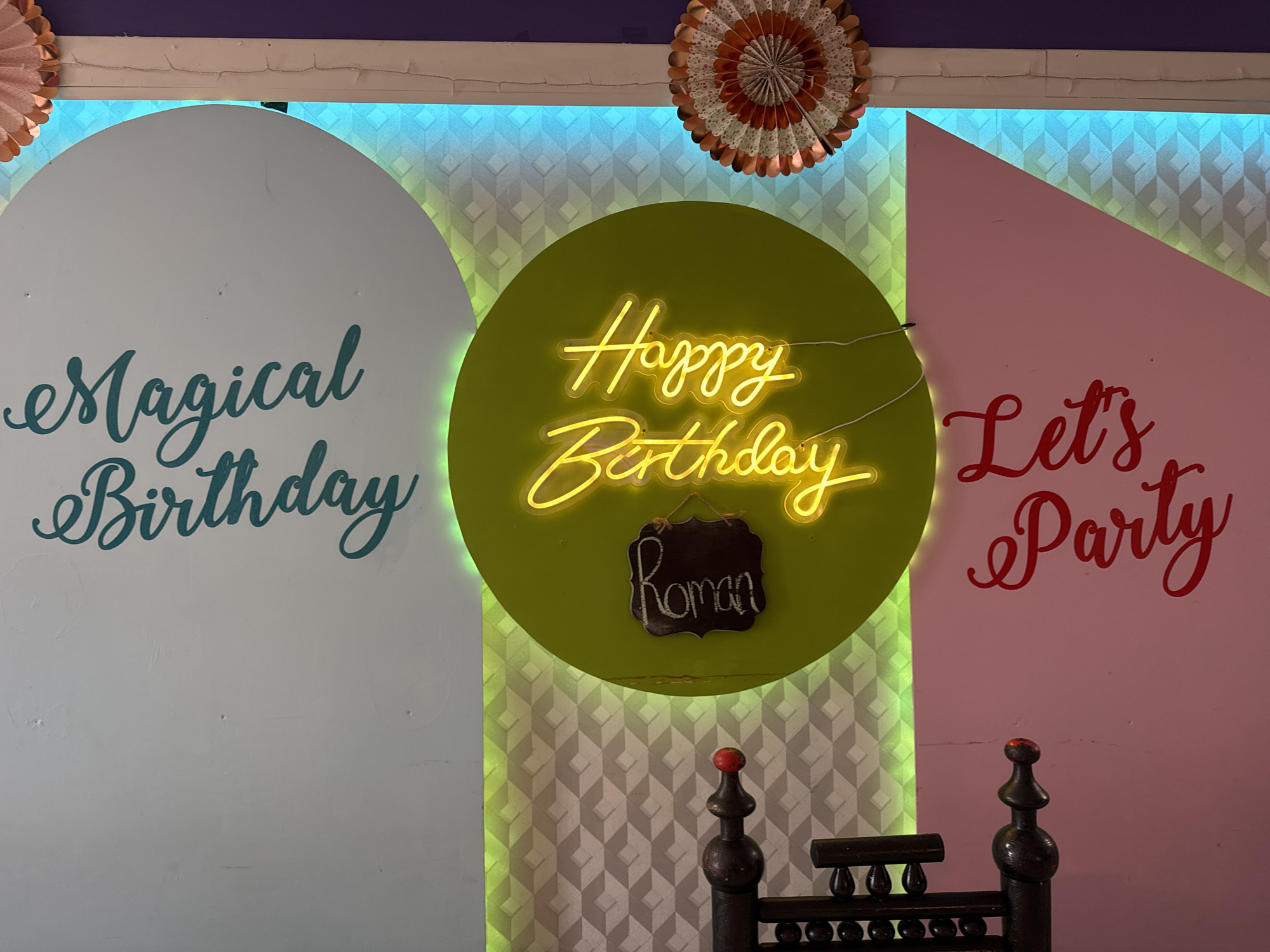 The image depicts a brightly decorated wall with neon signs and text. On the left, in teal script, reads "Magical Birthday". In the center, on a green circular background, a yellow neon sign says "Happy Birthday" with a small chalkboard sign below it that reads "Roman". To the right, in red script, reads "Let's Party". The wall behind the signs has a geometric patterned wallpaper illuminated with blue and green lights. Decorative paper fans are visible at the top of the image. The lower portion of the image shows part of a dark wooden chair with decorative finials. The scene appears to be a festive backdrop, possibly for a party or event, located in Clonmel, Ireland. There are no people visible in the image.