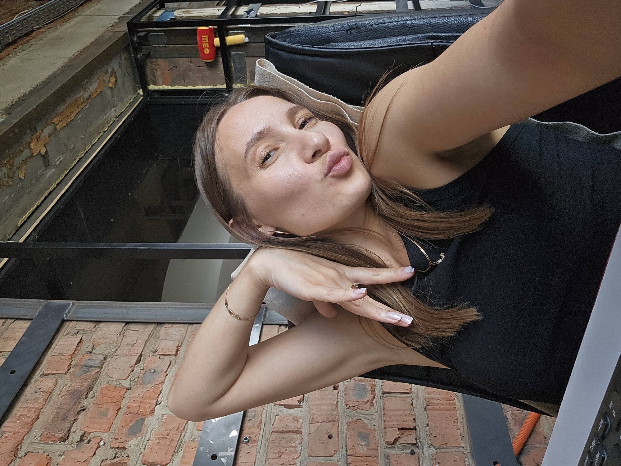 A woman is taking a selfie in an indoor setting that appears to be an office or industrial space. She is seated and leaning back in a black leather chair, with her left arm raised and bent behind her head. Her right hand is resting on her neck, with fingers splayed. She is wearing a black tank top and a gold bracelet on her left wrist, and a gold ring on her right index finger. She has long brown hair, and is wearing hoop earrings. Her lips are pursed as if she is about to kiss the camera. Behind her, a brick wall is visible, along with metal framing and some exposed wiring. A yellow and red object resembling a toy hammer hangs on a shelf in the background. The image is well-lit, with natural light appearing to come from the right. The location is not specified as Chaikovsky, Russia.