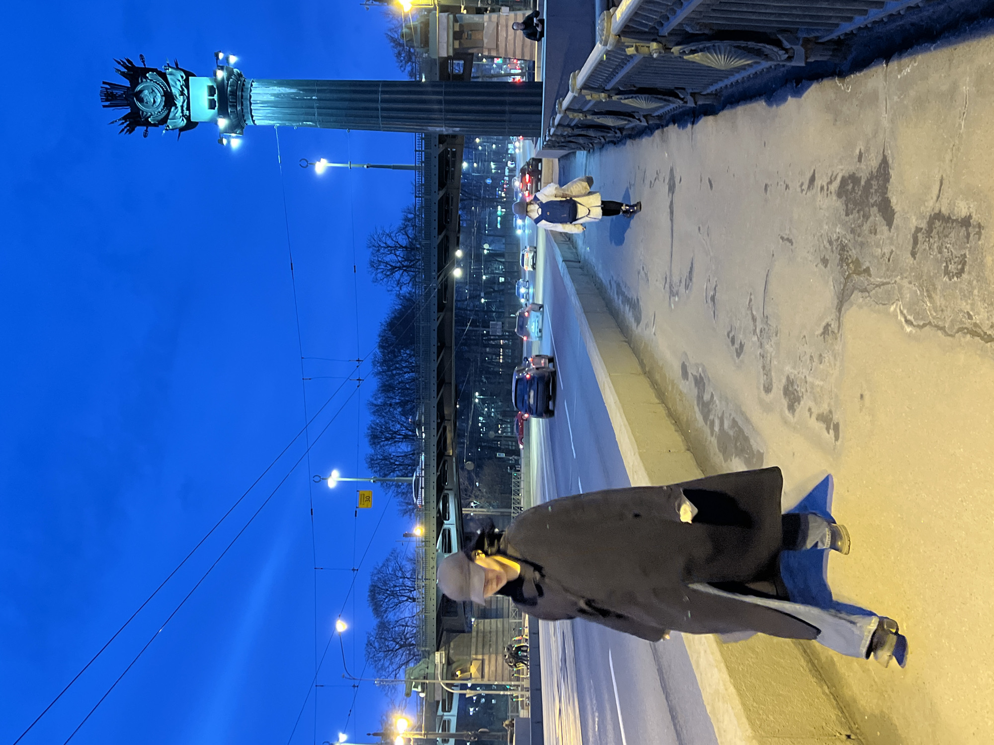 The image depicts an urban evening scene on Vasilyevsky Island, Saint Petersburg, Russia, near one of the Rostral Columns, with the Exchange Bridge (Birzhevoy Most) visible in the background. The deep blue sky indicates dusk or night.
In the foreground, a person walks towards the viewer on the left side of the frame, positioned on a concrete sidewalk. This individual wears a dark long overcoat, blue jeans, dark shoes, and a dark baseball cap that obscures their face. A yellow object is visible around their neck/ear area. To their left is a low concrete barrier separating the sidewalk from a multi-lane roadway.
Further down the same sidewalk on the right, a second person walks away from the viewer. This individual wears a light-colored coat, dark pants, dark boots, and a grey hat. They carry a dark blue backpack and a light-colored bag in their right hand. A white face mask is visible on the back of their head.
The roadway to the left of the sidewalk shows moderate vehicle traffic, with numerous cars displaying illuminated headlights and taillights. In the mid-ground, the ornate, dark metallic structure of the Exchange Bridge is visible, spanning horizontally across the scene with vehicles moving beneath and on it.
Prominently on the far right, a large, dark fluted Rostral Column stands tall, illuminated with a blue-green light, highlighting its intricate sculptural crest at the top, which features an imperial emblem and anchors. Several white lights are mounted on the column's base, casting upward illumination. Another person is dimly visible standing or sitting near the base of this column on the far right.
The sidewalk surface shows patches of moisture or ice, reflecting the artificial light from numerous streetlights that cast a yellow-orange glow on parts of the scene. Overhead wires, likely for trams or trolleybuses, traverse the sky. A yellow diamond-shaped traffic sign with "30" is visible on a streetlight pole in the background. Bare trees line the far background, completing the urban landscape.