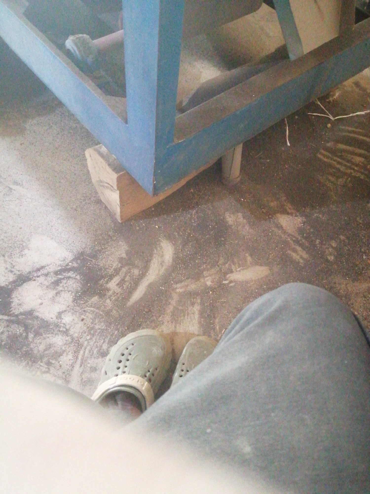 In the city of Jos, Nigeria, a low-angle shot captures a close-up view of a person's feet, clad in light green perforated clogs, resting on a dusty concrete floor. The person is wearing loose-fitting grey pants. In the background, a portion of a blue metal structure, possibly a table or support, is visible. A piece of light-colored wood is placed beneath the blue structure. The floor is unevenly covered with dust and debris, suggesting an outdoor or workshop environment. The lighting is moderate, with some areas appearing brighter than others. The image appears to be taken indoors or in a sheltered outdoor space.