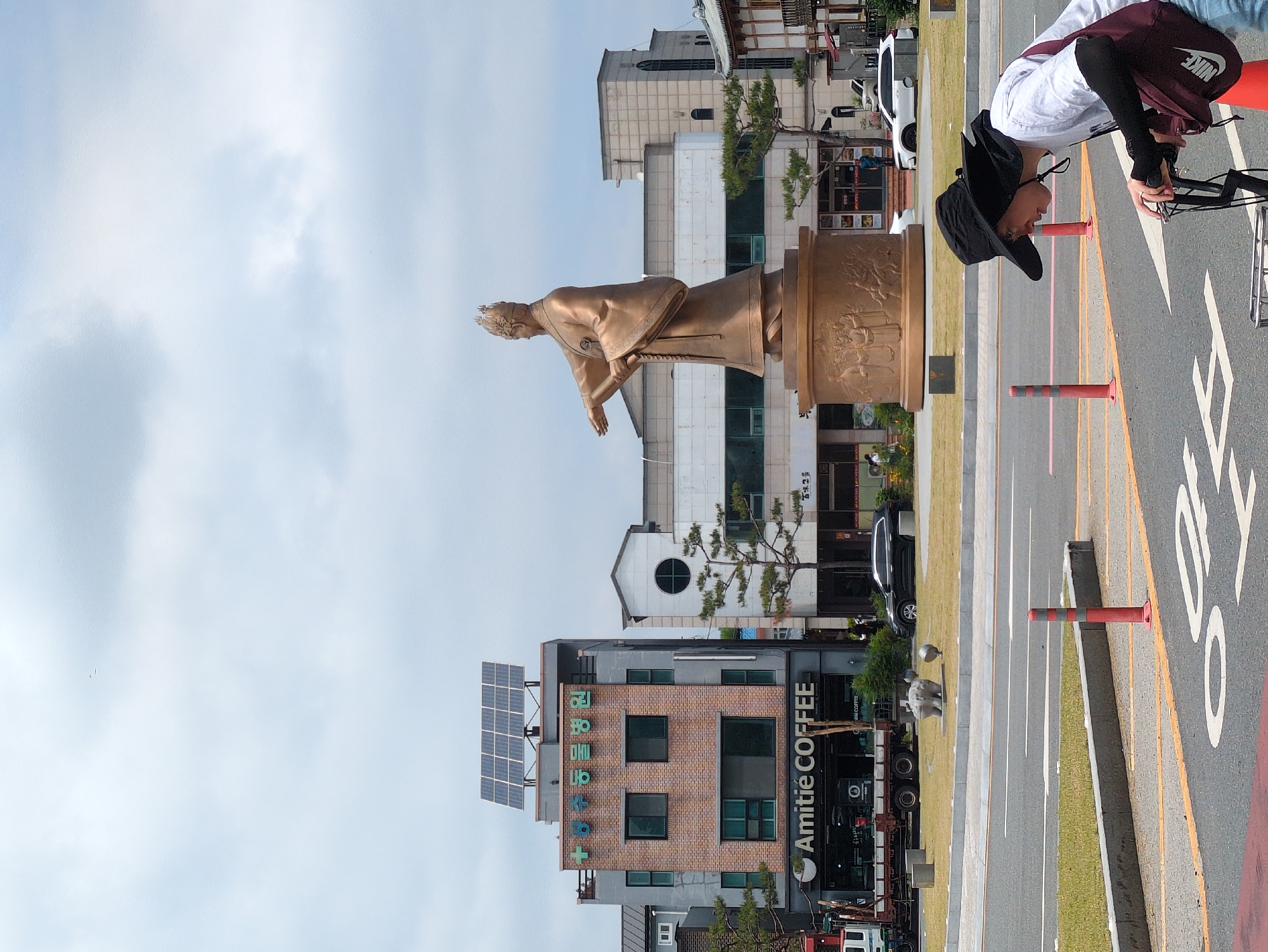 The image shows a bronze statue of a person with outstretched arms in Gongju-si, South Korea. The statue is mounted on a tall base with sculpted figures around it. The buildings in the background vary in height, materials, and architectural styles. One building has solar panels on its roof, and the sign "Amitie COFFEE" is visible. The sky is cloudy. The person in the image is wearing a large brimmed hat, a white shirt and a maroon/black bag is riding a bike. There are orange and grey striped bollards alongside the road and markings on the pavement.