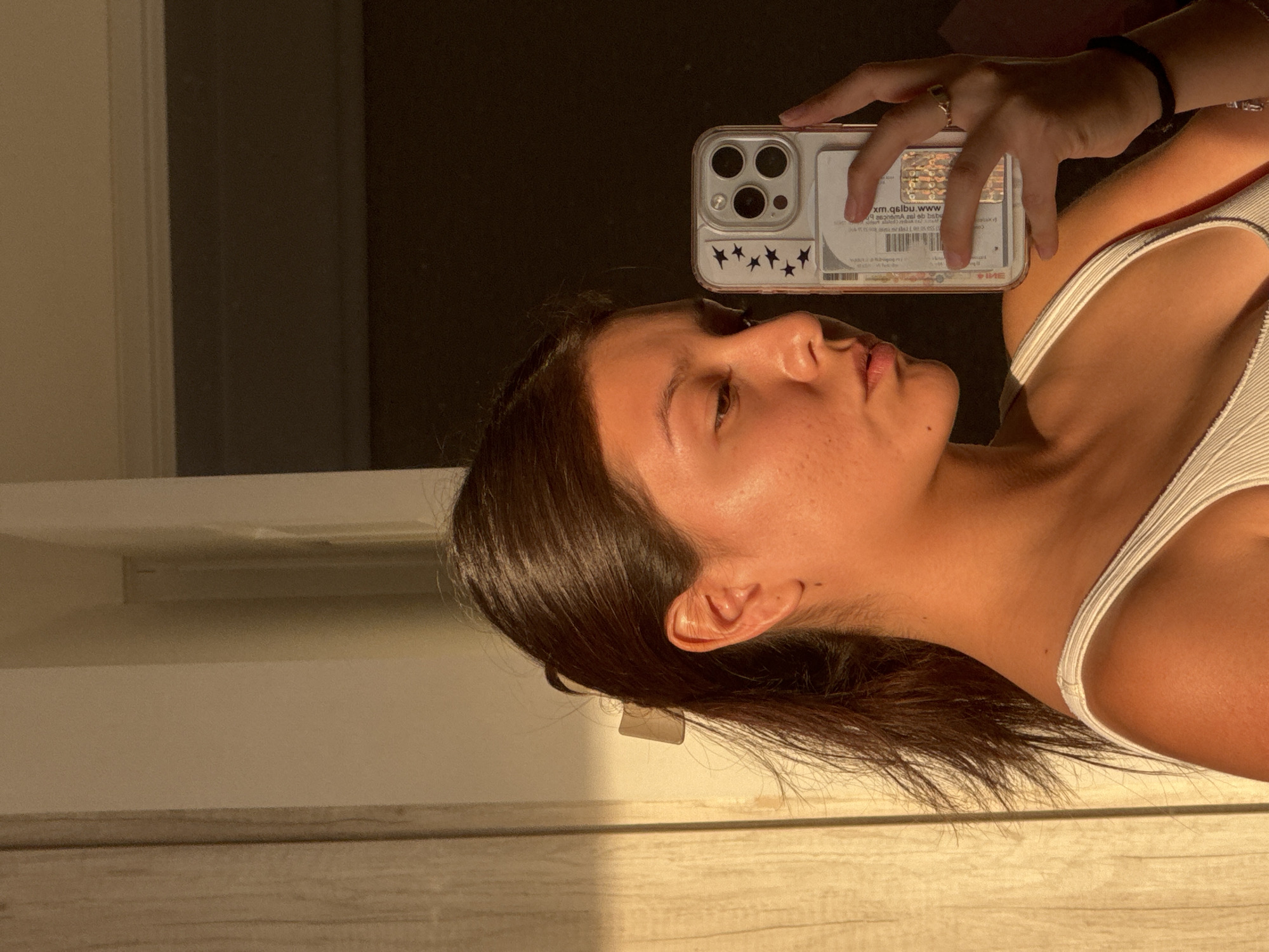 A reflective selfie captures a young woman in an indoor setting. Her face is presented in profile, looking slightly upwards and to the side, with her dark hair cascading around her neck and shoulder. She wears a white ribbed tank top.
She is holding a smartphone with a clear case that features small dark star stickers. Inside the phone case, a card is visible with mirrored text and a QR code. Partially legible mirrored text includes "ww.qslbu.xm" and "www." She also wears a ring on her finger and a black elastic band on her wrist.
The scene is illuminated by strong, warm, natural light, likely sunlight, which casts golden tones on her skin and creates prominent shadows. The background consists of varying shades, from very dark areas to lighter walls or surfaces, with a light-colored, possibly wooden, surface in the foreground. Her expression is calm, with lips slightly parted. The visual cues suggest the photograph was taken during the "golden hour" of early morning or late afternoon. The location context is San Francisco Ocotlán, Mexico.