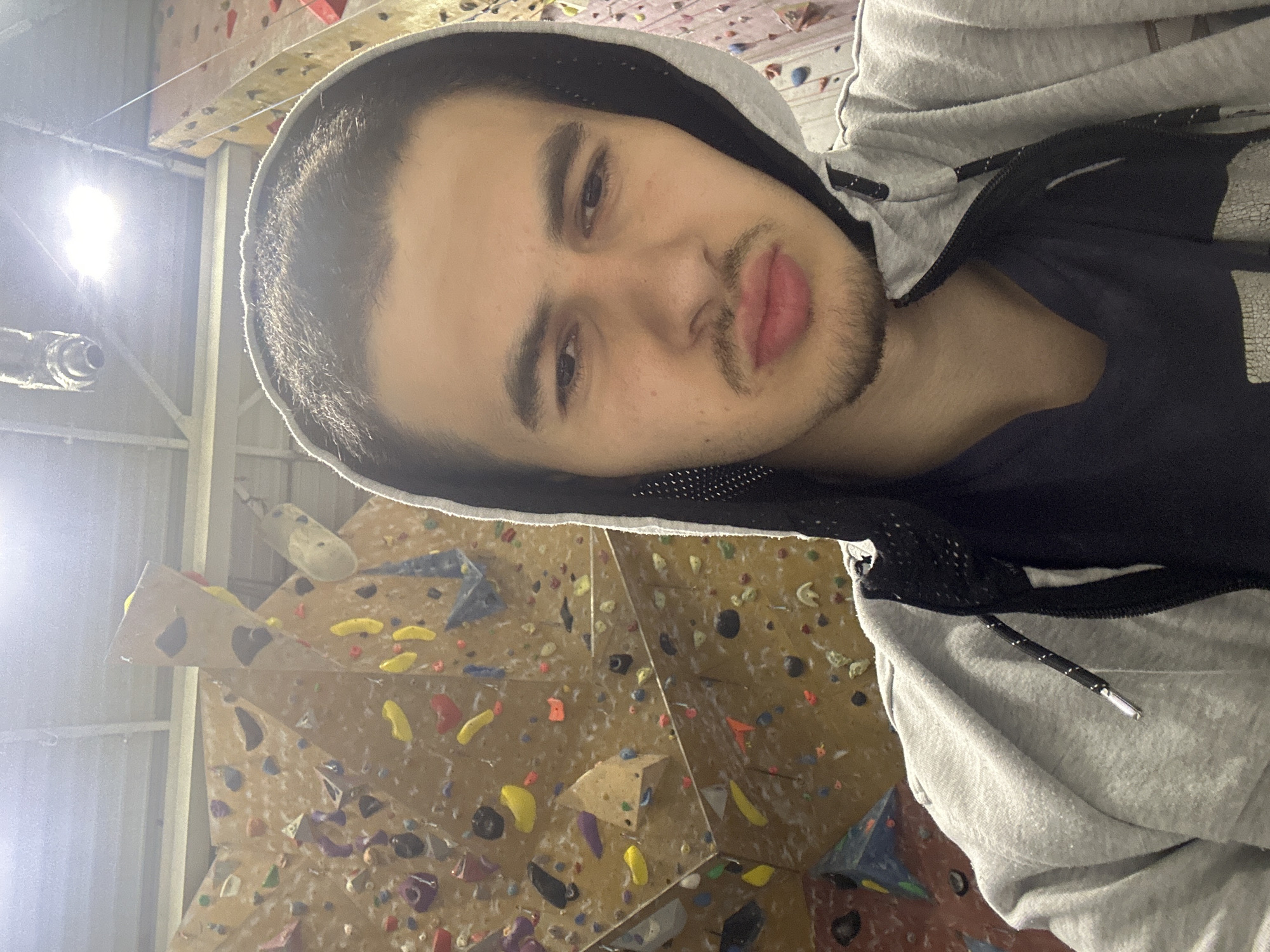 A selfie-style photograph shows a young man with dark hair and a slight beard wearing a gray hooded sweatshirt. The background reveals a climbing wall with numerous colorful holds. The photograph appears to have been taken indoors, likely in a climbing gym, but information about the location in Sofia, Bulgaria, is not discernible from the image.