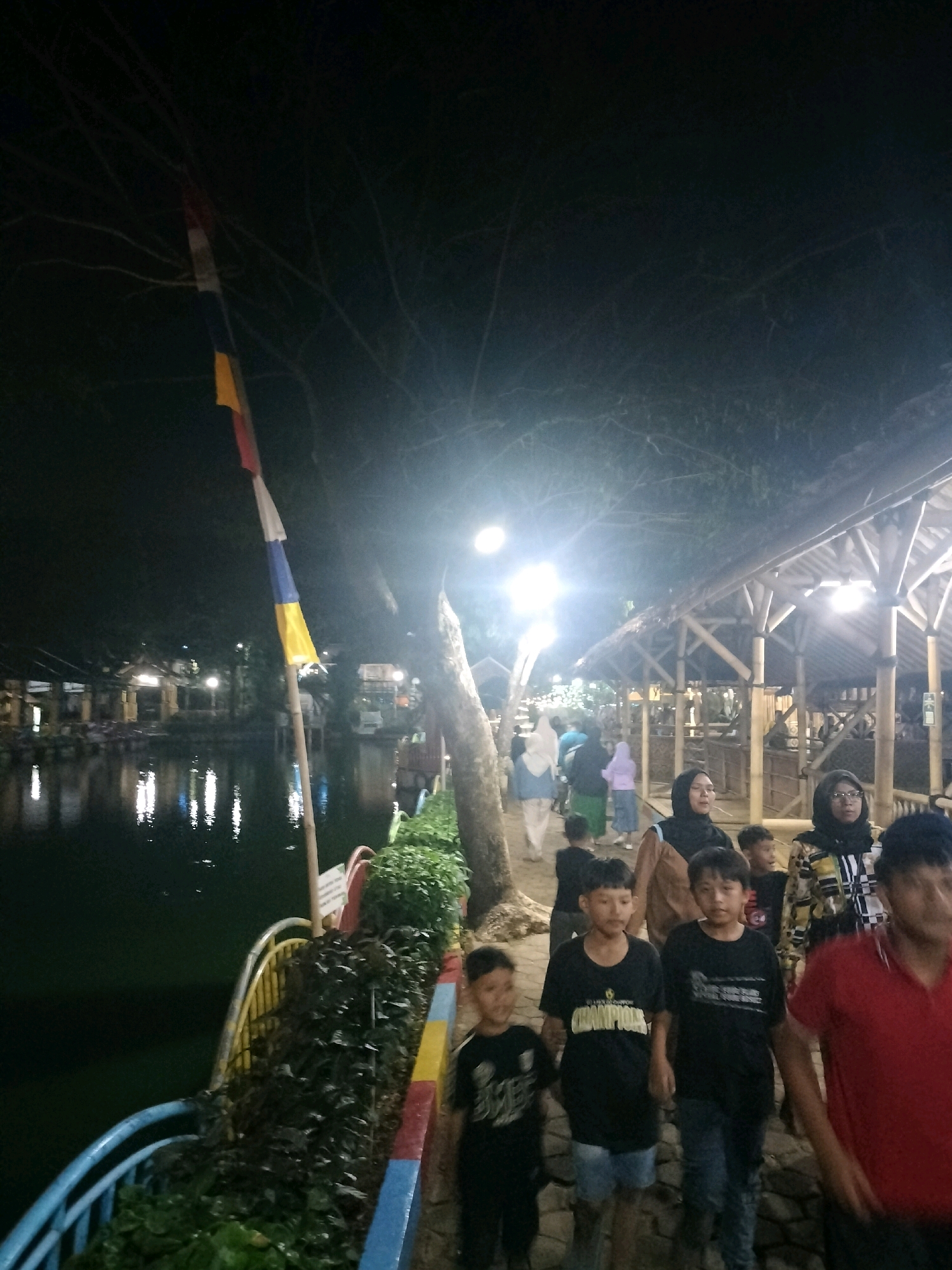 The image depicts an outdoor scene at night, likely a recreational area by a body of water, possibly in Depok, Indonesia, given the context of other similar images. In the foreground, a group of people are walking along a paved path. The individuals include several children and adults. Some of the children are wearing t-shirts with graphics. An adult male in a red shirt is partially visible on the right side of the frame. Behind them, to the left, is a dark body of water with reflections of lights from structures along its edge. These structures appear to be covered walkways or eateries with bamboo-like construction.
In the mid-ground, more people are visible, some dressed in headscarves. The path is lined with lush green vegetation and a colorful railing. A tall bamboo pole with horizontally striped flags in red, white, blue, and yellow is prominent on the left side. The background is dimly lit, with several bright lights illuminating the area, suggesting a festive or commercial atmosphere. The overall impression is of a lively gathering place during an evening outing.