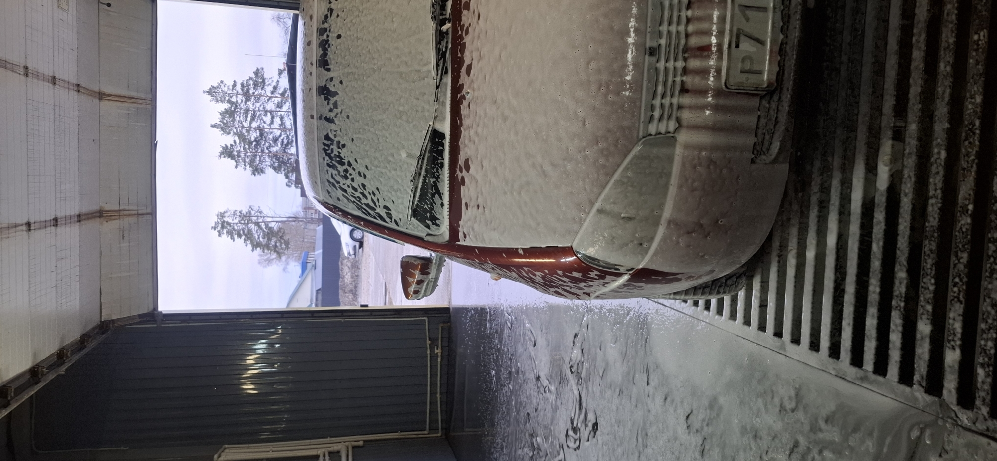 A red car is being washed with soap in a car wash in Chaikovsky, Russia. The car is covered in white foam. The license plate is visible. The car is parked over a grate. Through the open doorway, trees can be seen. The inside of the car wash is a dark gray color.