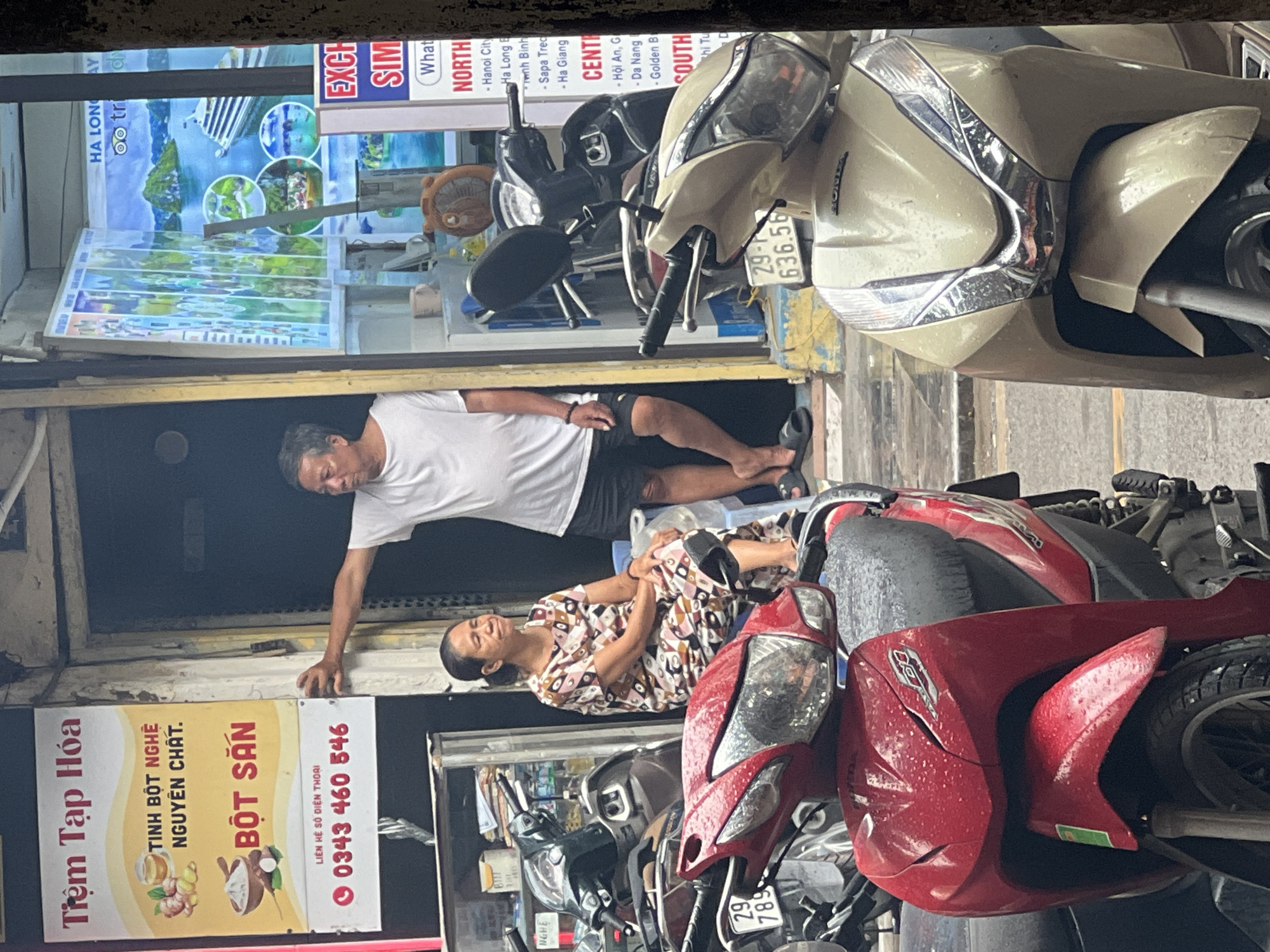 The image captures a street scene in Hà Nội, Vietnam, featuring a storefront with an advertisement and two individuals. A man in a white t-shirt and black shorts stands partially in a doorway, leaning against the frame. To his left, a woman in a patterned dress sits on a blue chair, her gaze directed upwards and to the left. In the foreground, several motorcycles are parked, with two prominently displayed: one red and one cream-colored. The storefront displays a sign with Vietnamese text and images, and further inside, another sign advertises travel destinations with images of Ha Long Bay. The environment appears to be a bustling urban street, with reflections on the wet surfaces of the motorcycles suggesting recent rain.