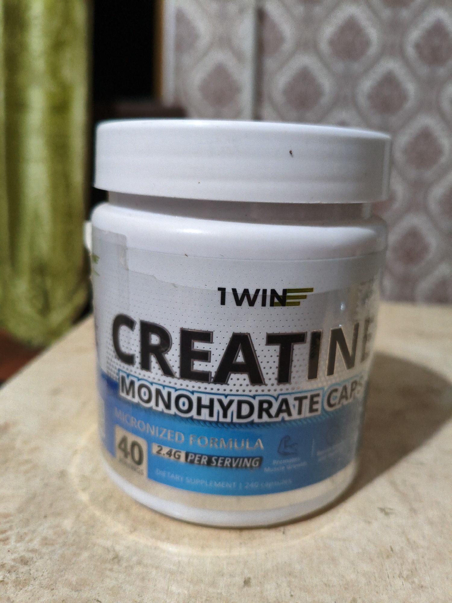 A white plastic bottle of "CREATINE MONOHYDRATE CAPS" by "1 WIN" sits on a wooden surface. The bottle has a white lid and a label with black text and blue accents. The label indicates "MICRONIZED FORMULA," "2.4G PER SERVING," and "40 SERVINGS." In the background, a green ribbed object is visible on the left, and a wall with a diamond pattern is on the right. The scene is indoors, on a table or counter.