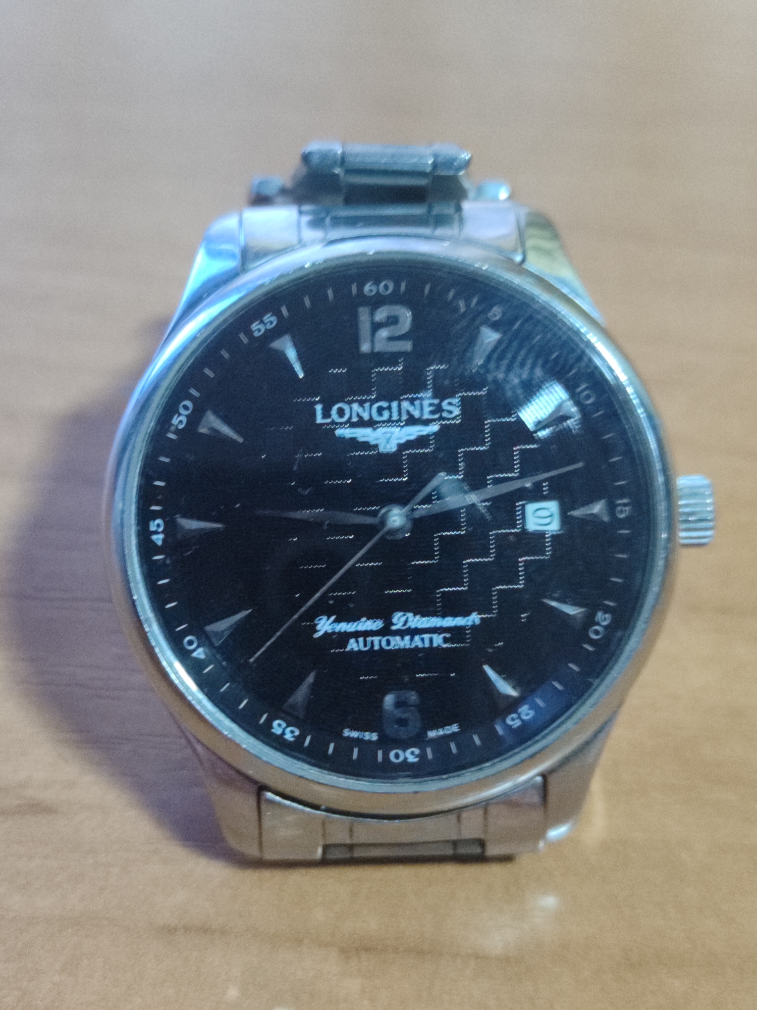 A close-up, eye-level, slightly tilted shot shows a stainless steel Longines wristwatch with a black face and a diamond pattern. The watch has silver hour markers and hands, with the number "12" prominently displayed at the top. Text on the face reads "LONGINES," "Genuine Diamonds," and "AUTOMATIC." The bezel is marked with numbers from 5 to 60 in increments of 5. The watch is resting on a wooden surface.
