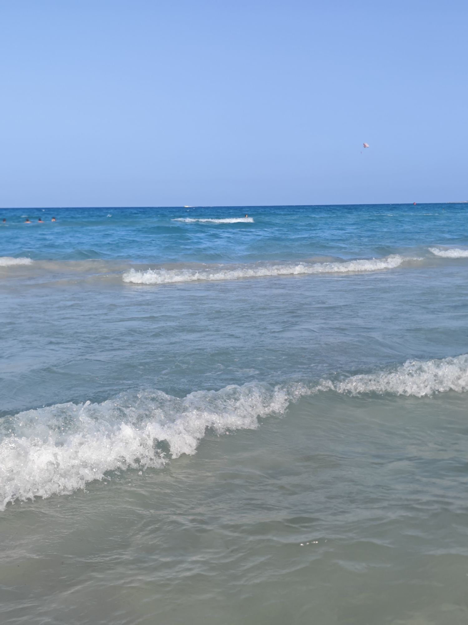 The image shows a view of the sea in Mahdia, Tunisia, under a clear blue sky. Gentle waves are breaking on the shore, creating white foam. In the distance, several people can be seen swimming or wading in the turquoise water. A red buoy is visible near the horizon, and a small white boat or structure can also be spotted further out. A colorful kite is flying high in the sky, adding a touch of activity to the serene scene. The water is calm with a few ripples and the sea bed is visible through the clear water near the shore.