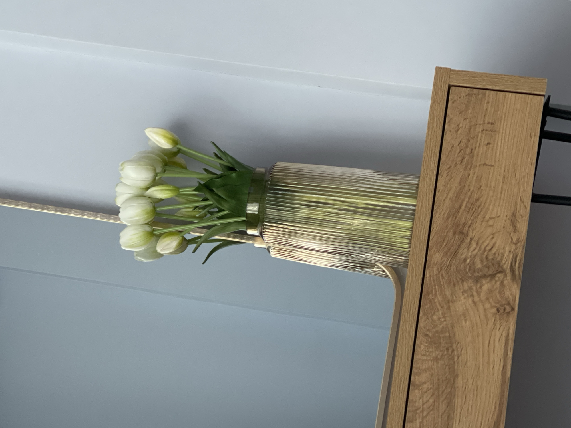 A close-up, indoor shot shows a bunch of white tulips in a ribbed, golden-toned glass vase. The vase is situated on a wooden surface, possibly a shelf or the top of a drawer, which has a visible wood grain texture. Behind the vase and tulips, a light blue wall meets a white wall, with a subtle vertical line where they join. The foreground is dominated by the wooden surface and the vase.