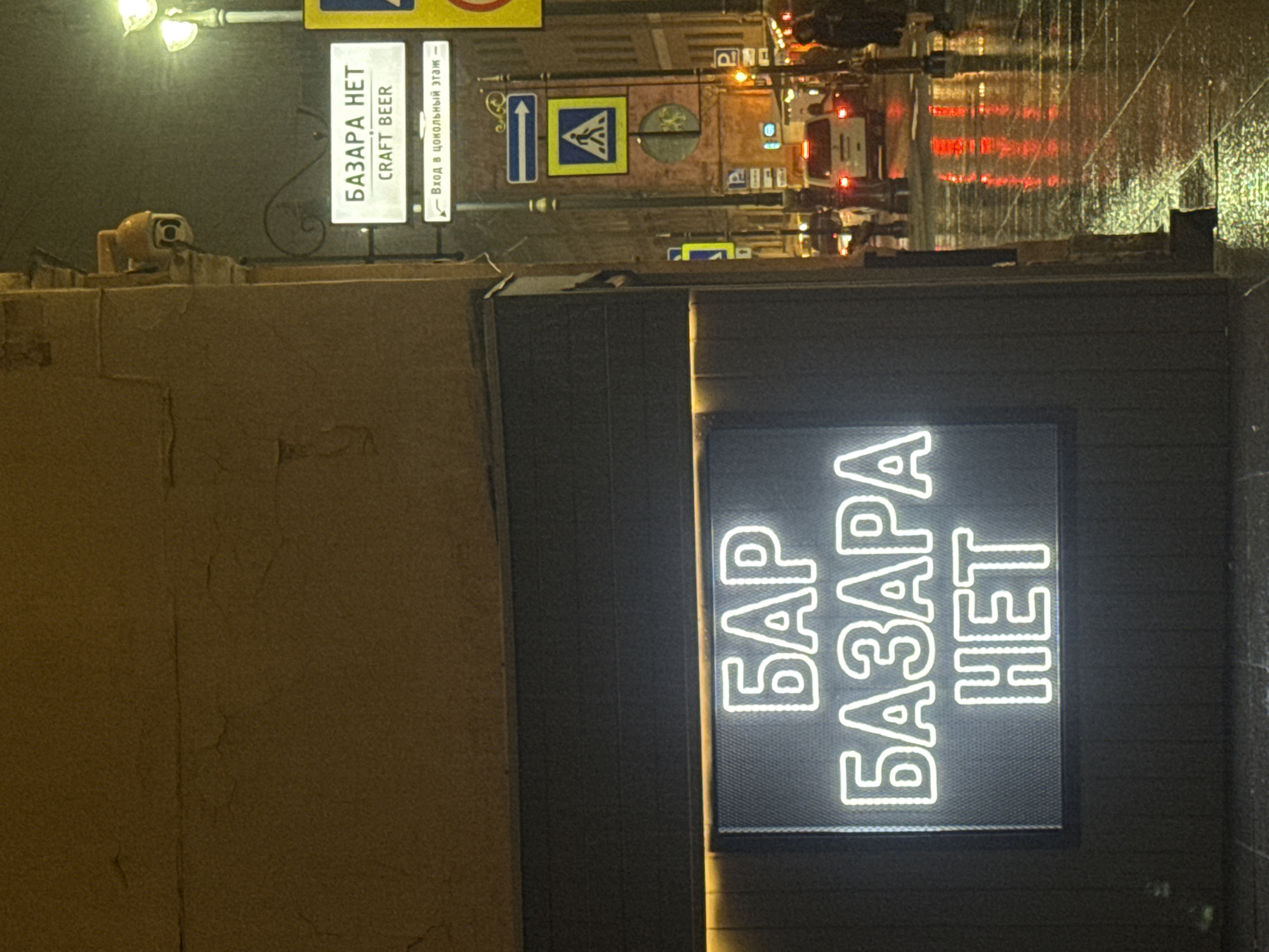 This nighttime image captures a street scene in Saint Petersburg, Russia. The illuminated sign of a bar, "БАР БАЗАРА НЕТ" (BAR BAZARA NET), is prominently displayed on the right, featuring white, glowing block letters against a dark background. Adjacent to this, a weathered, beige wall extends to the left. Above, a street lamp casts light on a series of signs, including one with "БАЗАРА НЕТ CRAFT BEER" (BAZARA NET CRAFT BEER), as well as traffic signs, directional arrows, and parking symbols. In the background, a street is visible with reflections from the wet pavement, suggesting rain. Cars with illuminated headlights and taillights are present, along with pedestrians. The overall impression is that of a city street at night.