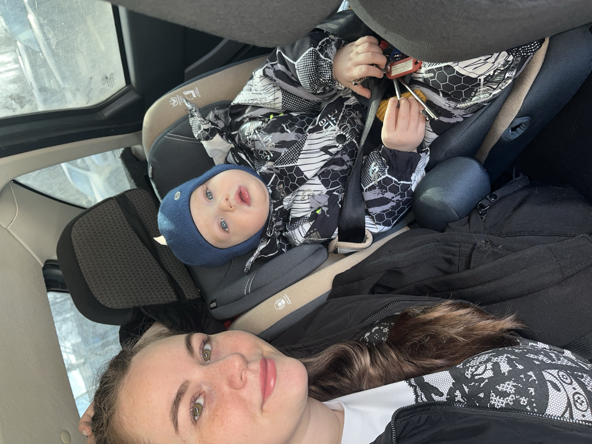 This image shows two individuals inside a car in the city of Chaikovsky, Russia. A young adult female is positioned in the foreground on the left, capturing a selfie. She has long, wavy dark brown hair, fair skin with visible freckles, green eyes, and light pink lips. She wears a black zippered jacket over a white top with a black lace-like pattern and looks directly at the camera with a slight smile.
To her right and slightly behind her, an infant is secured in a grey and beige child safety seat, held by a harness system. The infant, with light blue eyes, wears a dark blue knit beanie and a black, white, and grey patterned winter suit featuring hexagonal and abstract designs. A small amount of clear liquid is visible on the infant's lower lip. The infant holds a set of keys with a red key fob in their right hand and looks slightly to the right of the camera.
The car's interior features dark grey fabric upholstery. Through the vehicle's windows, a blurred outdoor environment is visible, including trees or branches and other parked vehicles, but no specific landmarks.