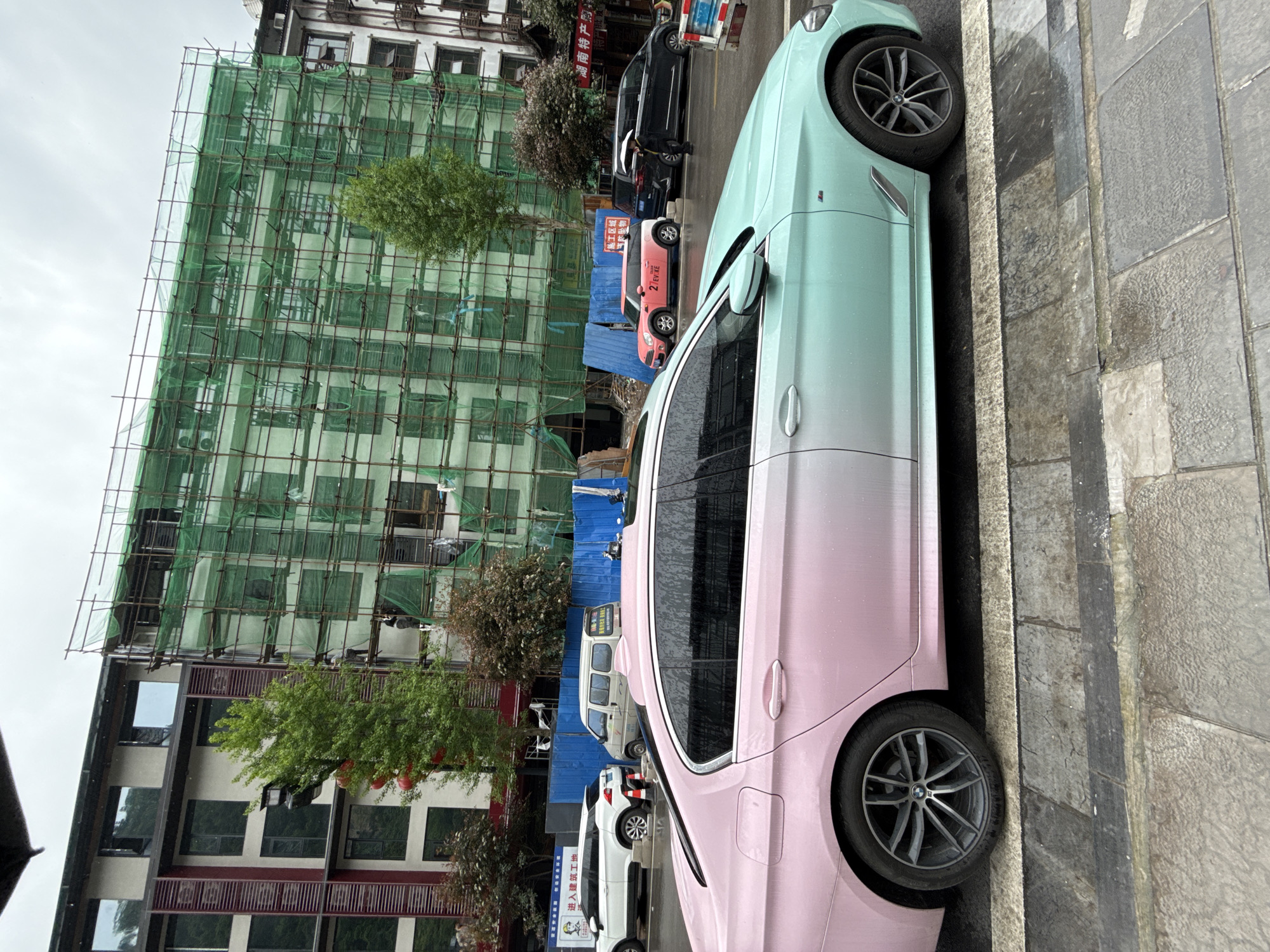The image, oriented horizontally, depicts a vibrant street scene in what appears to be an urban area of Wulingyuan, China. The primary subject is a uniquely painted car, a sedan, parked on the right side of the frame. Its exterior is split into two distinct colors: the rear half is a striking light pink, and the front half is a contrasting light teal or mint green. The car's wheels feature a BMW logo.
In the background, a large multi-story building is under extensive construction or renovation, enveloped in green scaffolding and netting. Scattered trees with green foliage are visible, one appearing to grow through the scaffolding. To the left of the construction site, another building with multiple windows and dark framing is visible. Further down the street, blue corrugated metal barriers mark the perimeter of a construction zone.
Several other vehicles are parked or driving in the background: a black SUV or van, a smaller orange car with the text "2° FIRE", a white van, and another white SUV. A person holding an umbrella can be faintly seen near the black car, suggesting recent rain or an overcast day. The street and sidewalk appear damp, reinforcing the impression of wet weather.
Visible Chinese text provides specific location cues: a distant sign reads "湖南特产店" (Hunan Specialty Store), and signs near the construction barriers state "施工区域 禁止入内" (Construction area, no entry) and "进入建筑工地" (Enter construction site). The sky is overcast, indicating daytime. The scene captures an ordinary moment of urban life amidst development and construction.