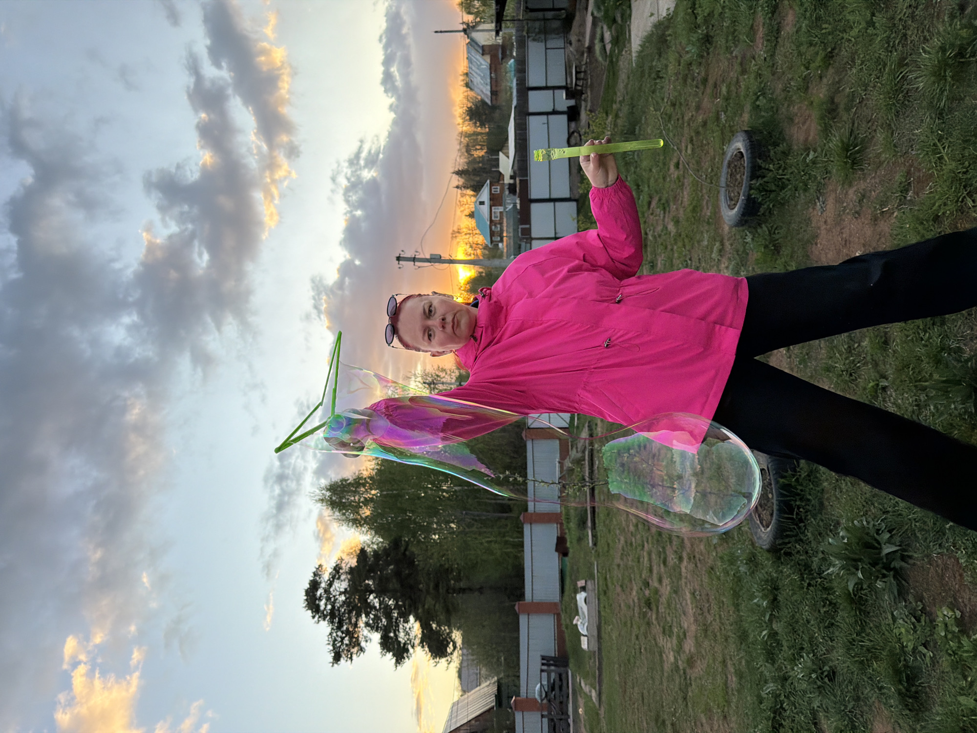 A woman with glasses on her head wears a pink jacket and black leggings, standing in a grassy yard under a sky with sunset colors. She is blowing giant bubbles with a green bubble wand. The background includes a fence, trees, and houses. The location is Chaikovsky, Russia.