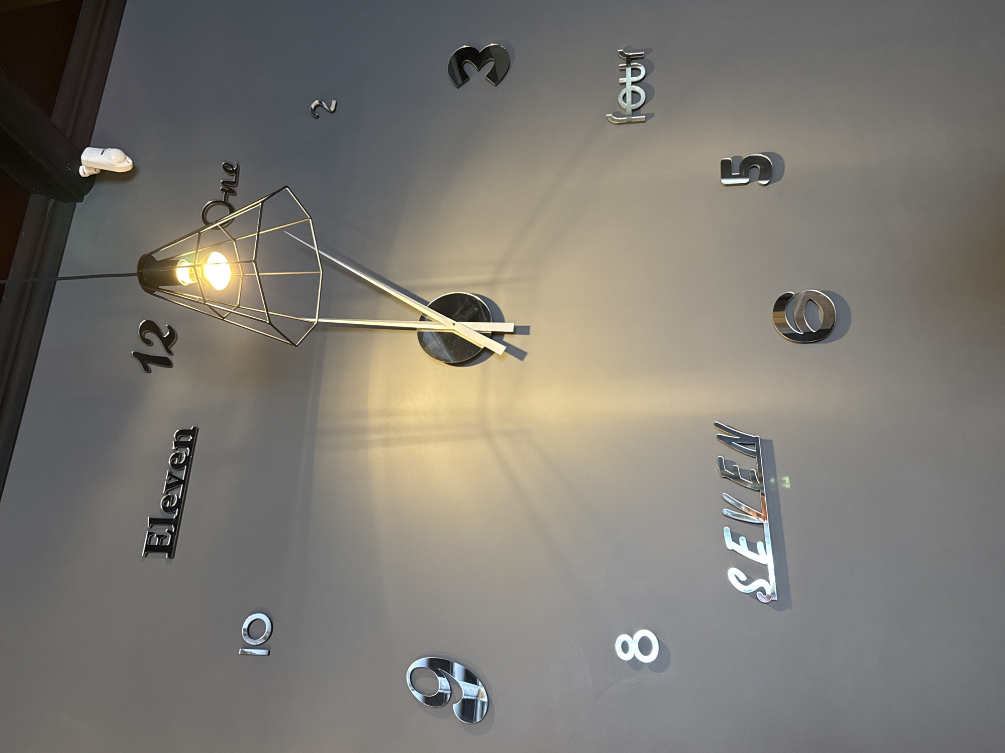 This image depicts a large, modern wall clock. The clock face is composed of metallic 3D numbers and words representing hours. The numbers visible are: '12', '1', '2', '3', '4' (written as "four"), '5', '6', '7' (written as "SEVEN"), '8', '9', '10', and 'Eleven'. A central mechanism with clock hands is also present, positioned near the '12' and '1'. Above the clock, a pendant light with a wire cage design hangs down, casting a warm glow on the wall. A small white motion sensor is mounted high on the wall to the right of the light. The background is a plain, gray wall, suggesting an indoor setting. The image was taken in the city of Kirishi, Russia.