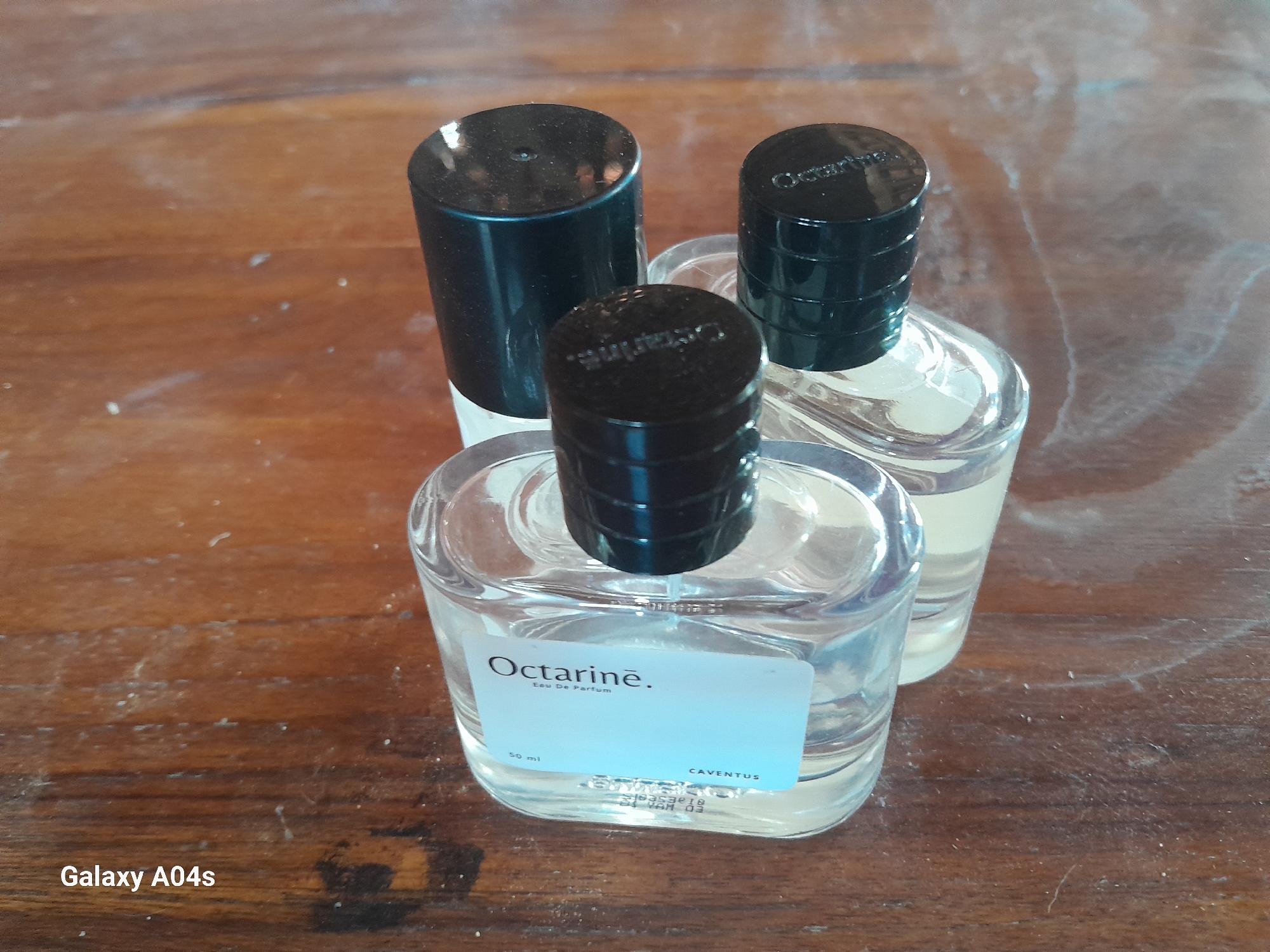 Three glass perfume bottles with black caps are placed on a wooden surface. The bottle in the foreground has a white label that reads "Octarine. Eau De Parfum 50 ml CAVENTUS". The other two bottles, positioned behind the first, also have black caps and clear glass bodies, with a faint label visible on the middle bottle's cap that appears to read "Octarine". The wooden surface has visible wood grain and some scattered white dust or particles. The bottom left corner of the image displays the text "Galaxy A04s". The scene is indoors, with the lighting suggesting it is daytime. There are no people or animals visible in the image. The location is identified as Pilangpayung, Indonesia.