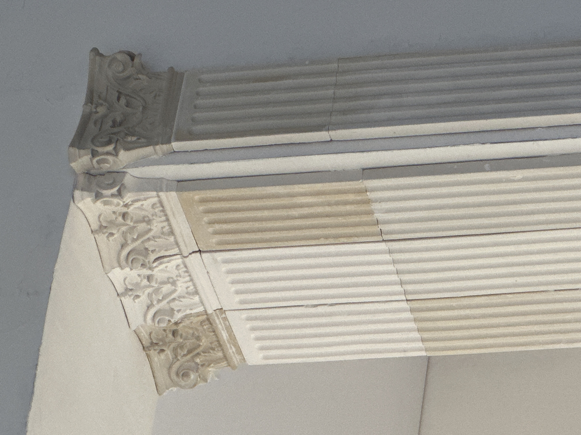 The image shows a close-up of architectural detailing, specifically a section of an entablature or cornice. The architectural elements are predominantly white or light-colored, possibly made of stone or plaster. The components include a decorative corner piece adorned with floral or scroll-like carvings, and horizontal bands with repeating parallel grooves or ridges. The structure is assembled from separate blocks or sections, visible through the seams between them. The image suggests an indoor location, likely within a building with classical or neoclassical design. Location: Unknown, Unknown.