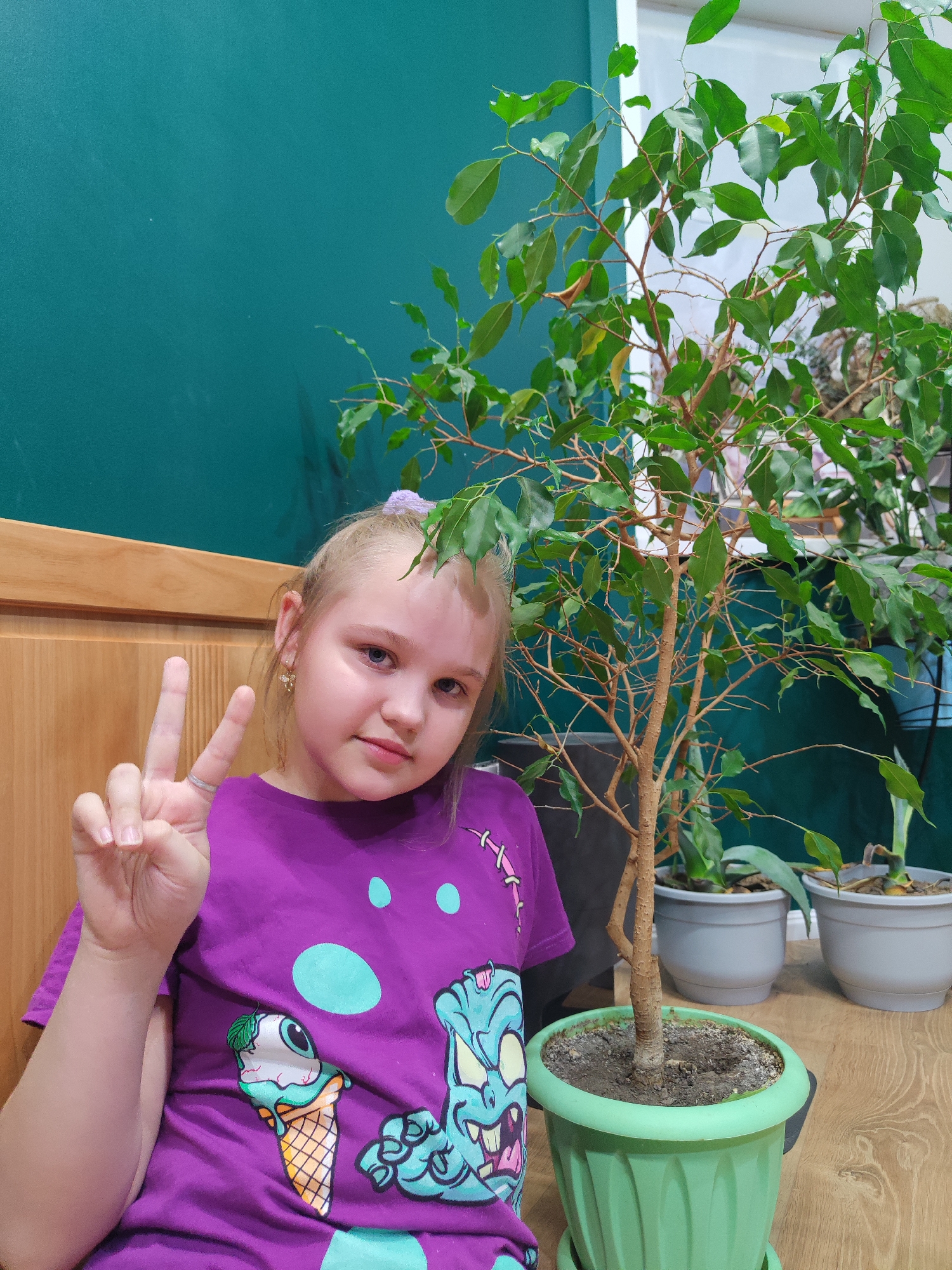 A young, light-skinned girl is sitting indoors near a wooden wall and a green wall. She is wearing a purple t-shirt with a cartoon monster and an ice cream cone graphic and has blonde hair pulled back with a light purple hair tie. She is holding up her hand in a peace sign. Next to her, there is a tall ficus tree in a green pot. Behind that tree are two more plants in grey pots. The scene is set indoors, likely in a house or apartment. Based on the request, the location is Chaikovsky, Russia.