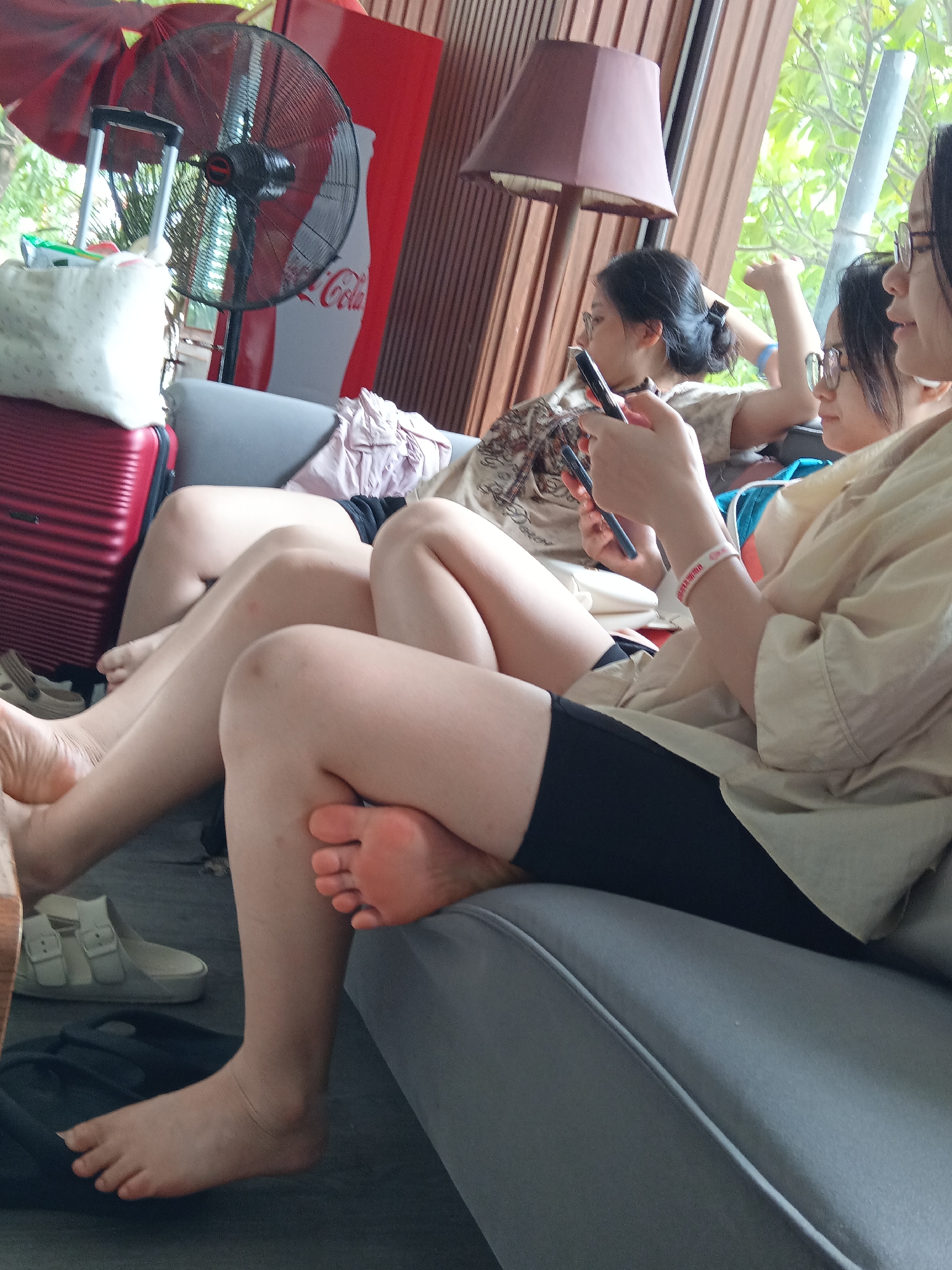 The image depicts three young women relaxing on a gray sofa. The woman on the left is wearing a light-colored shirt with a patterned trim, and black shorts. She is holding a smartphone in her left hand, with her legs crossed. The woman in the middle is wearing glasses and a light-colored shirt. She is also holding a smartphone and appears to be interacting with it. Her legs are bent at the knees. The woman on the right, also wearing glasses, is wearing a light-colored button-up shirt and is looking towards the left. Her arm is extended, and she is holding a smartphone. The background shows a Coca-Cola branded fan, a dark-colored lamp with a brown shade, and wooden paneling. The lighting suggests an indoor setting, possibly a cafe or lounge. Given the presence of a Coca-Cola branded fan and the overall aesthetic, the location could be a tourist-oriented establishment in Đà Nẵng, Vietnam, such as a cafe near the beach or a relaxation area within a resort. A red suitcase and a white cloth bag are visible on the floor to the left. White sandals are also present near the suitcase. The environment appears casual and relaxed, with the women engrossed in their phones.