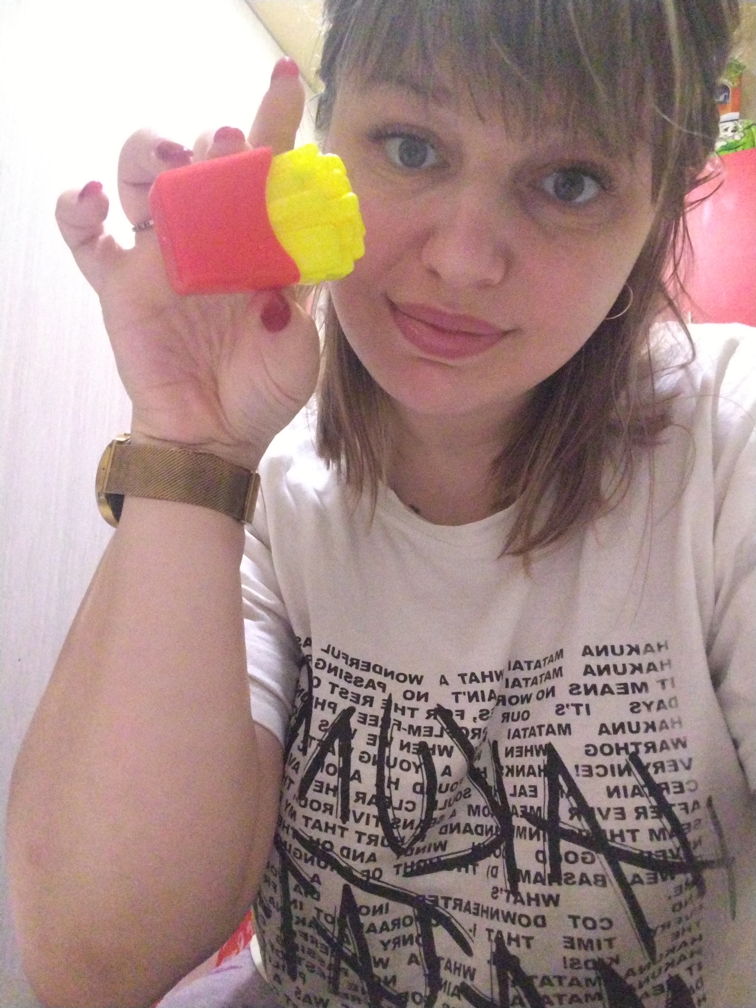 A fair-skinned woman with medium-length brown hair and bangs is holding a toy resembling a container of french fries in her left hand. The toy is red with yellow "fries" protruding from the top. She is wearing a gold-colored watch on her left wrist and a ring on her left hand. The woman is wearing a white t-shirt printed with black text and designs. The background appears to be a plain, light-colored wall. The location is unknown.