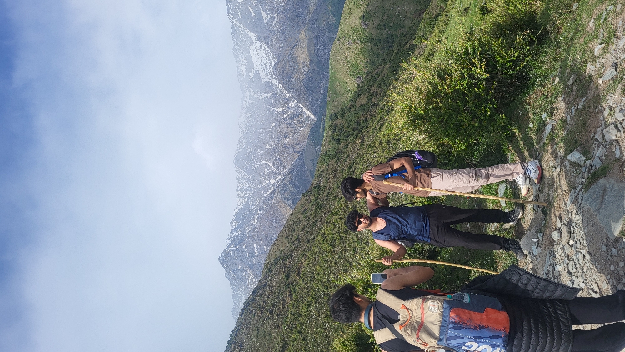 Three male subjects are positioned on a mountain hiking trail. Two of the subjects stand upright while one subject is hunched over holding a cellular phone. Each subject is carrying a backpack, and two of the subjects hold hiking sticks. The environment consists of rocky terrain, light green vegetation, and large snow-covered mountains in the distance. The time of day appears to be mid-day. The location is near Dharamshala, India.