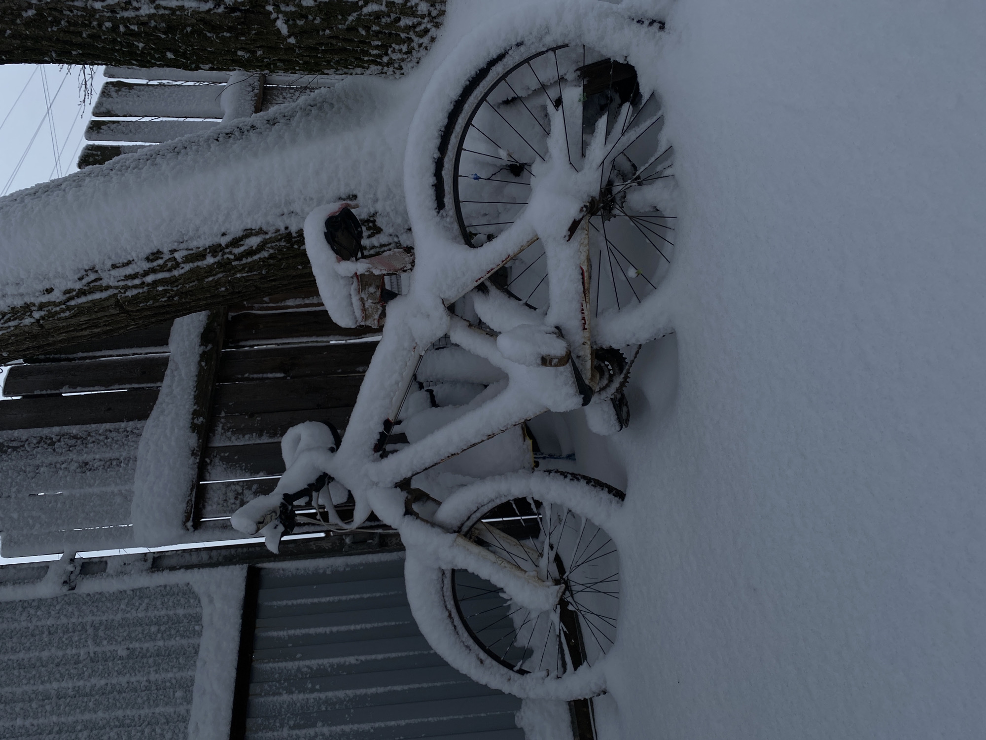 A bicycle is buried in snow, leaning against a wooden structure also covered in snow. The location appears to be outdoors with a fence-like structure visible in the background. The sky is overcast. The scene depicts a winter environment. The location is the city of Ольховка, Russia.