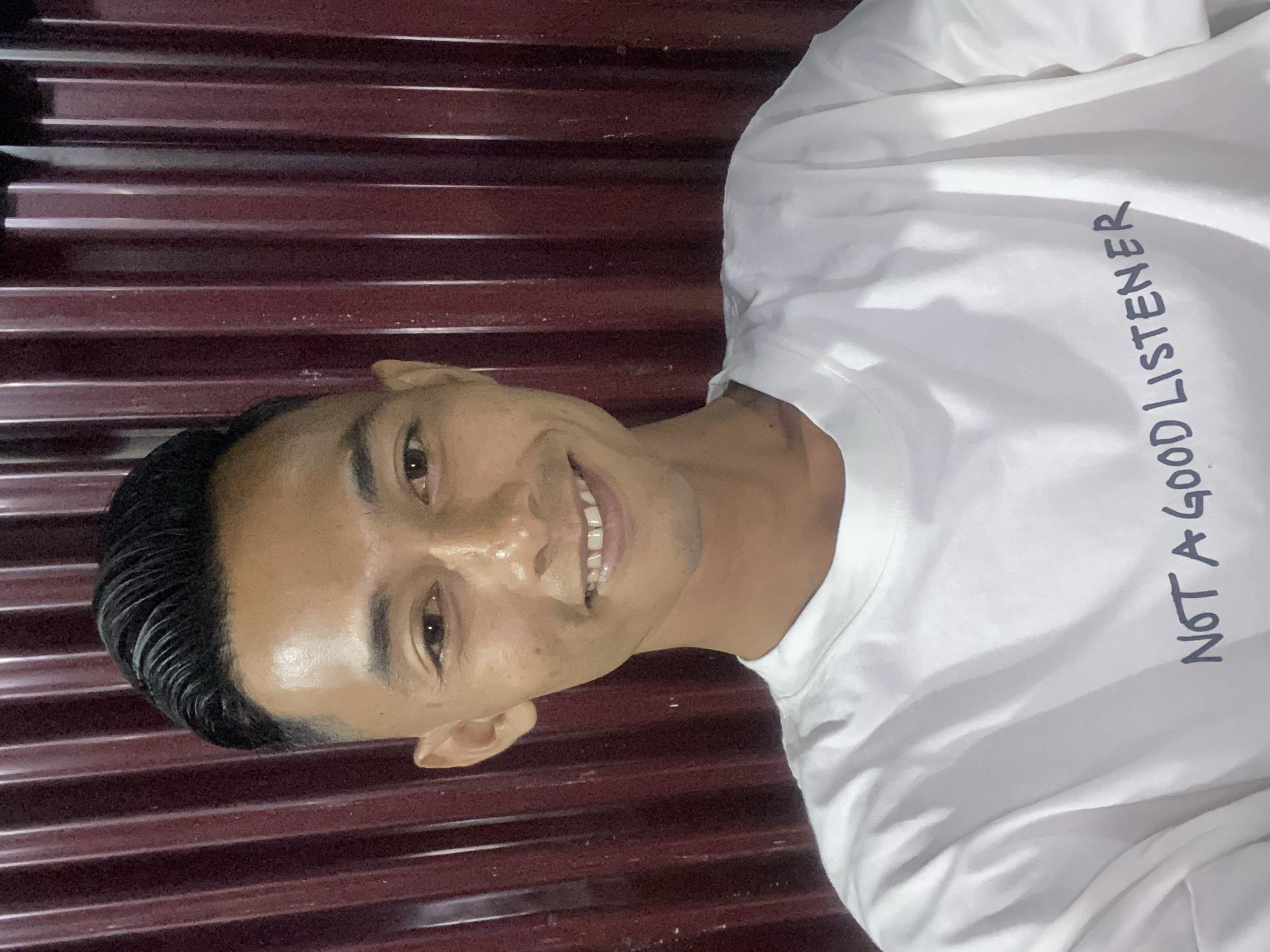 A young man is depicted in a close-up portrait, smiling broadly at the camera. He has short, dark, slicked-back hair and a cheerful expression, revealing his teeth. His skin has a slight shine, possibly from lighting or perspiration. He is wearing a plain white, long-sleeved t-shirt. Printed vertically on the front of his shirt, in dark, hand-written style capital letters, is the phrase "NOT A GOOD LISTENER". The background features a dark, reddish-brown corrugated surface, likely a metal wall or door, providing a textured backdrop with horizontal lines. The lighting appears direct, illuminating the man and his white shirt. The image captures a casual moment, with the subject conveying a friendly and approachable demeanor. While the specific location isn't visually evident, the provided context suggests Gampongaceh, Indonesia.