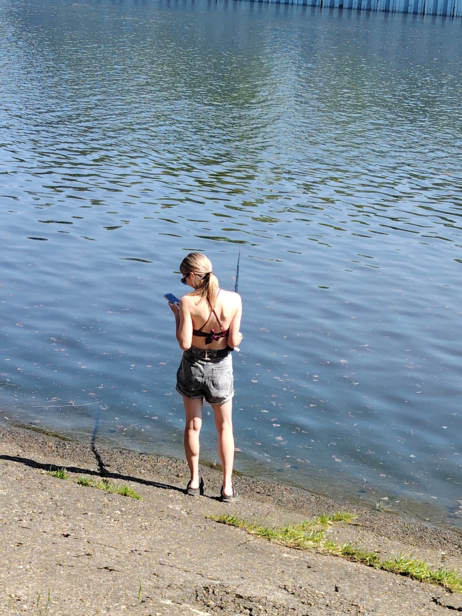 A woman stands facing a body of water, possibly the Kama River in Chaikovsky, Russia. She wears a dark-colored bikini top, grey denim shorts, and sandals. She is holding a fishing rod in her right hand and a smartphone in her left. The water is slightly rippled, and there are concrete embankments with grass lining the edge of the water. A distant structure appears across the water. The day is sunny.