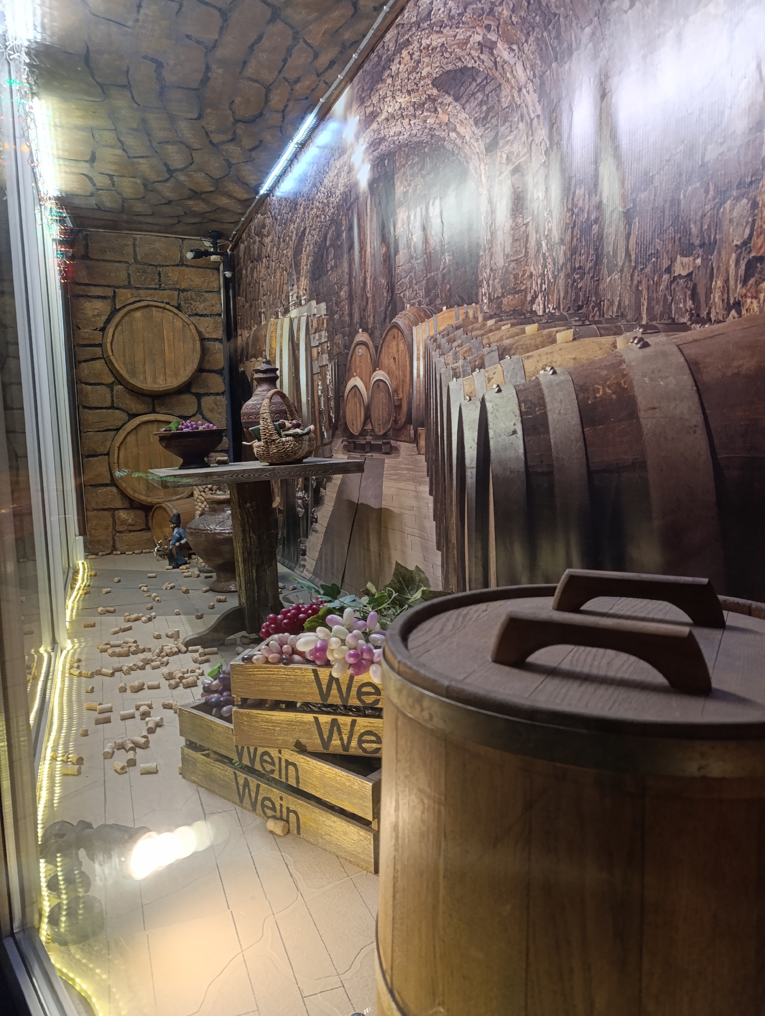 The image depicts a staged wine cellar display, possibly located in Chernogolovka, Russia. The scene is visible through what appears to be a glass window, indicated by light reflections.
The background simulates a stone cellar interior, with a brick-textured wall on the left and a backdrop depicting a wine cellar with rows of aging barrels.
In the foreground, a display table holds pottery and fruit. Wine corks are scattered on the floor around the table. In front of the table is a stack of wooden crates with "Wein" (German for wine) stenciled on them, overflowing with artificial grapes and leaves.
A large wooden barrel with a lid and handles dominates the lower right portion of the frame.
The overall impression is that this is a stylized, artificial environment intended to evoke a traditional wine cellar.