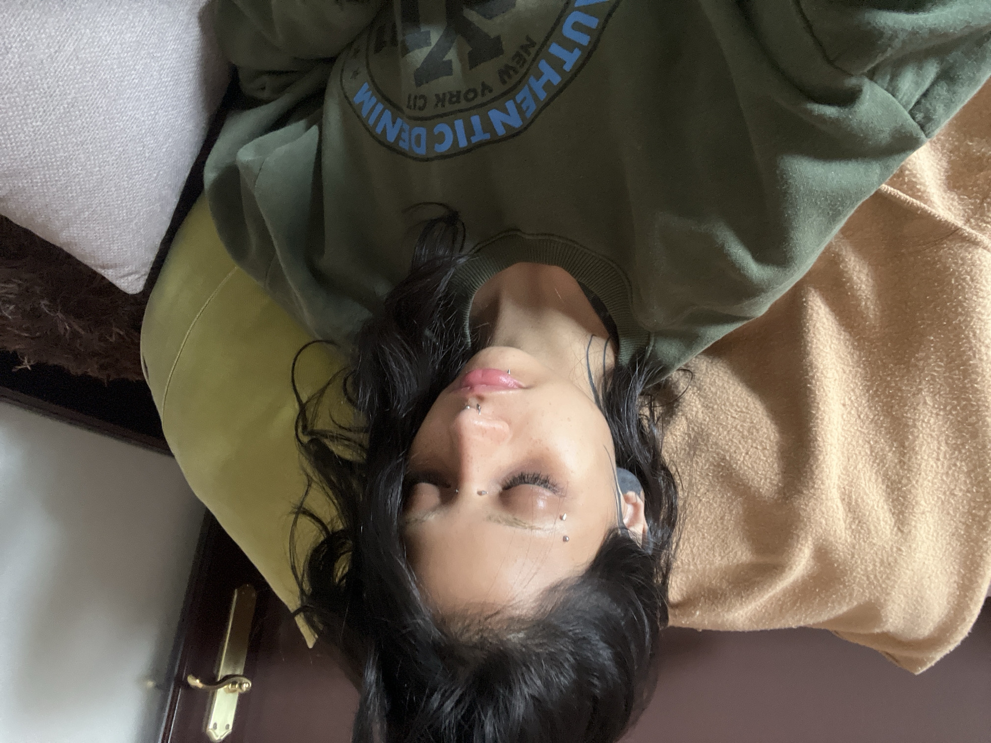 A woman is lying down, possibly on a bed or couch, wearing an olive green sweatshirt with the words "AUTHENTIC DENIM NEW YORK CITY" printed on it. Her dark hair is spread around her head. She has piercings on her face, including her lip and above her eyebrows. She is wearing earbuds in her ears. The background includes a white pillow, a yellow pillow, and a tan blanket. A dark-colored door with a gold handle is visible in the lower left corner.
The location in the city of Arroyo De La Miel-benalmadena Costa, Spain cannot be verified from this image.