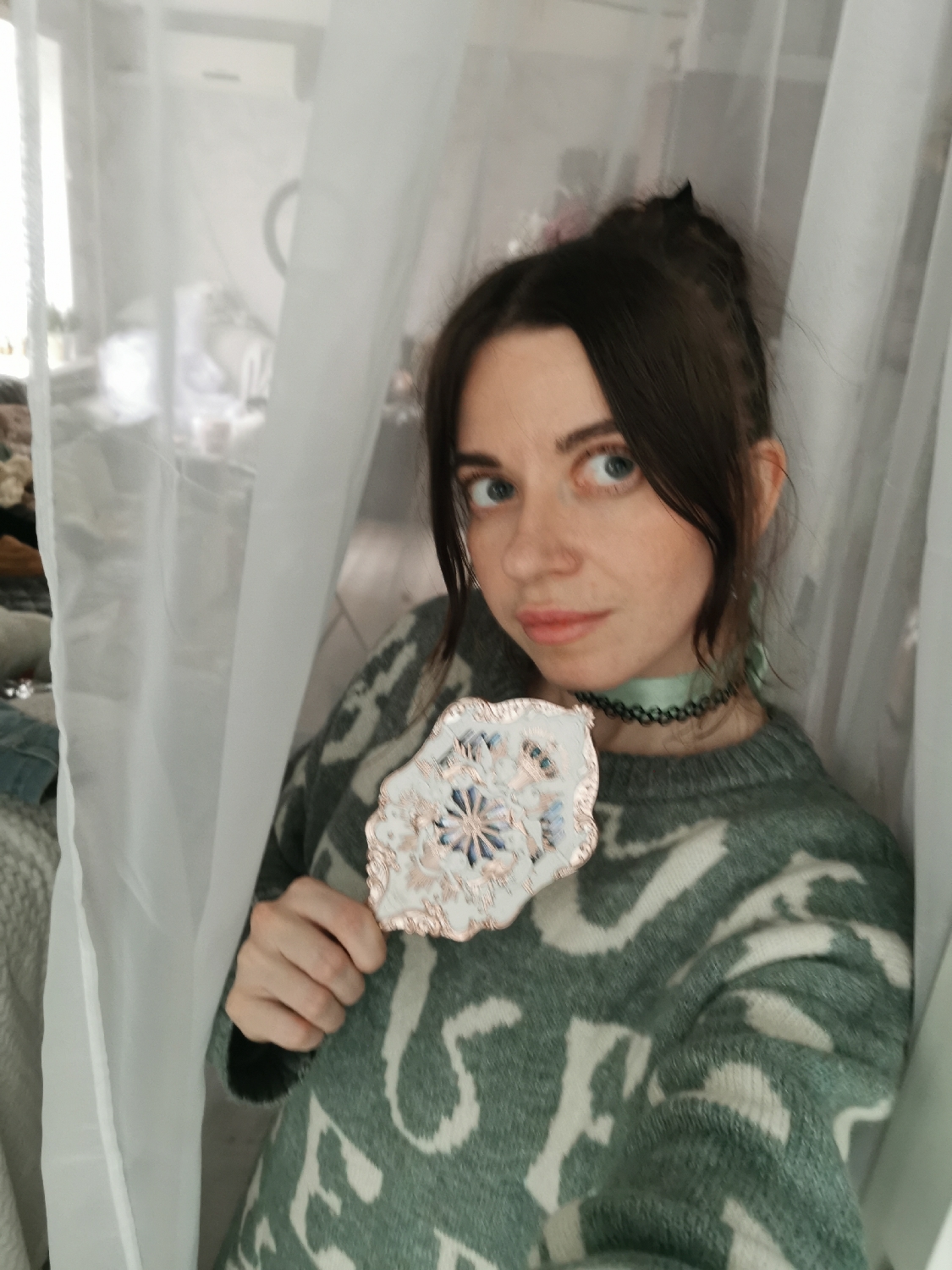 An indoor, eye-level photograph captures a woman framed by sheer white curtains. She has dark brown hair, pulled up into a bun with some strands framing her face, and light blue-green eyes. She wears a thick, green knitted sweater adorned with large, abstract off-white patterns, and a light green choker necklace.
In her right hand, she holds a decorative handheld mirror featuring an ornate, possibly rose gold-toned frame, and a white back embellished with a central blue and gold/rose gold design. The woman looks directly towards the camera with a neutral, slightly inquisitive expression.
The background is softly blurred and largely obscured by the white curtains, hinting at an indoor room, possibly a bedroom or living area. Diffused natural light suggests the photograph was taken during the daytime. No distinct, legible text is visible within the sweater's pattern.