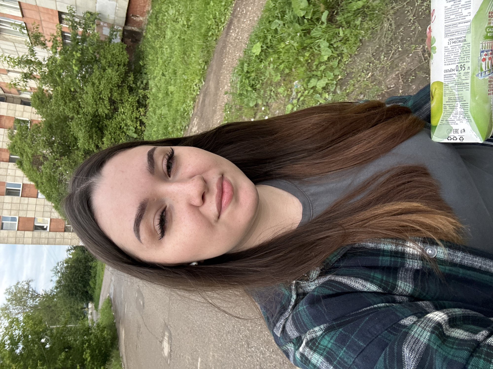The image shows a young woman with long brown hair taking a selfie outdoors. She is wearing a green and blue plaid shirt over a gray t-shirt. She has prominent eyelashes and eyebrows, and her lips are slightly parted. In her right hand, she holds a carton of apple juice. Behind her, there is a paved road, a grassy area with some plants, and a multi-story building with a reddish-brown facade and white window frames. The scene appears to be in a residential area, possibly in the city of Perm, Russia, given the context of the original request. The sky is overcast.