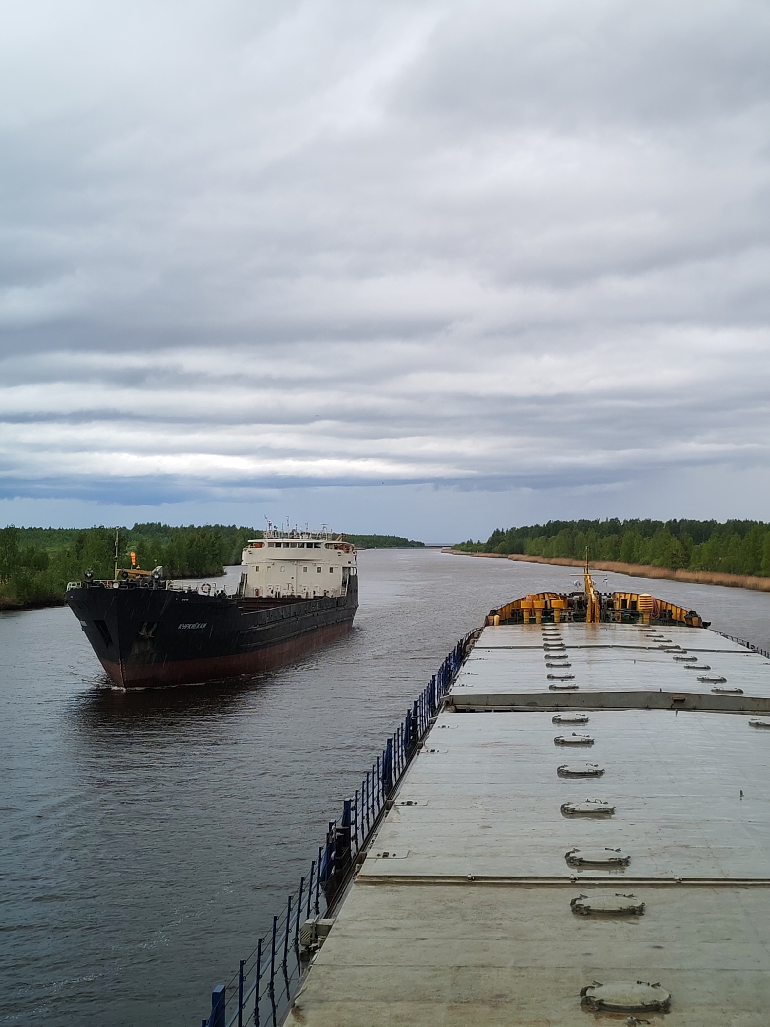 The image depicts two ships traversing a canal in Ozernoe Ust'e, Russia. The canal is lined with green trees on either side. The sky is overcast with gray clouds. One ship, visible in its entirety, is black and red with a white upper deck. The name "KУРИХЕCKИИ" is visible on its side. The second ship, occupying the foreground, appears to be a flat-deck barge or pontoon, with rows of hatch covers. The image is taken from the perspective of someone on the second ship looking towards the first. The water in the canal is dark and reflects the overcast sky.
