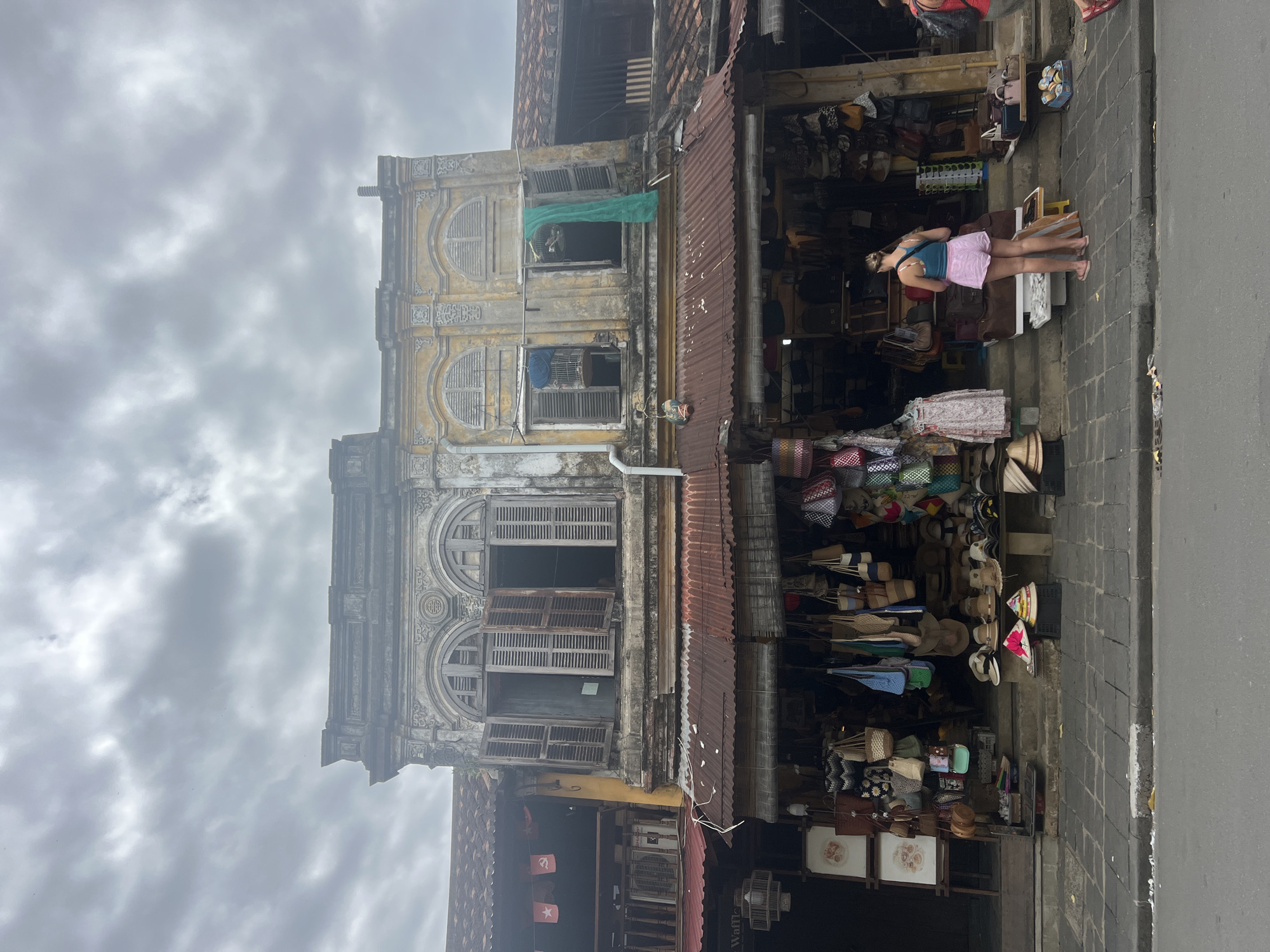 The image shows a street scene in Hoi An, Vietnam, with a focus on two adjacent buildings. The building on the left has a grey, ornate facade with arched windows that are boarded up. The building on the right is painted yellow and has similarly arched windows, with wooden shutters partially open. Both buildings appear aged and weathered.
In front of the buildings is a shopfront displaying a variety of goods, including bags, hats, and other accessories. The shop is covered by a rusty corrugated metal awning. A woman wearing a blue tank top and pink shorts stands facing the shop, looking at the merchandise. To the right of the woman, another person in a red shirt is partially visible walking away from the camera. The street in the foreground is paved with square tiles, and the sky is overcast. The overall atmosphere suggests a market or shopping area.