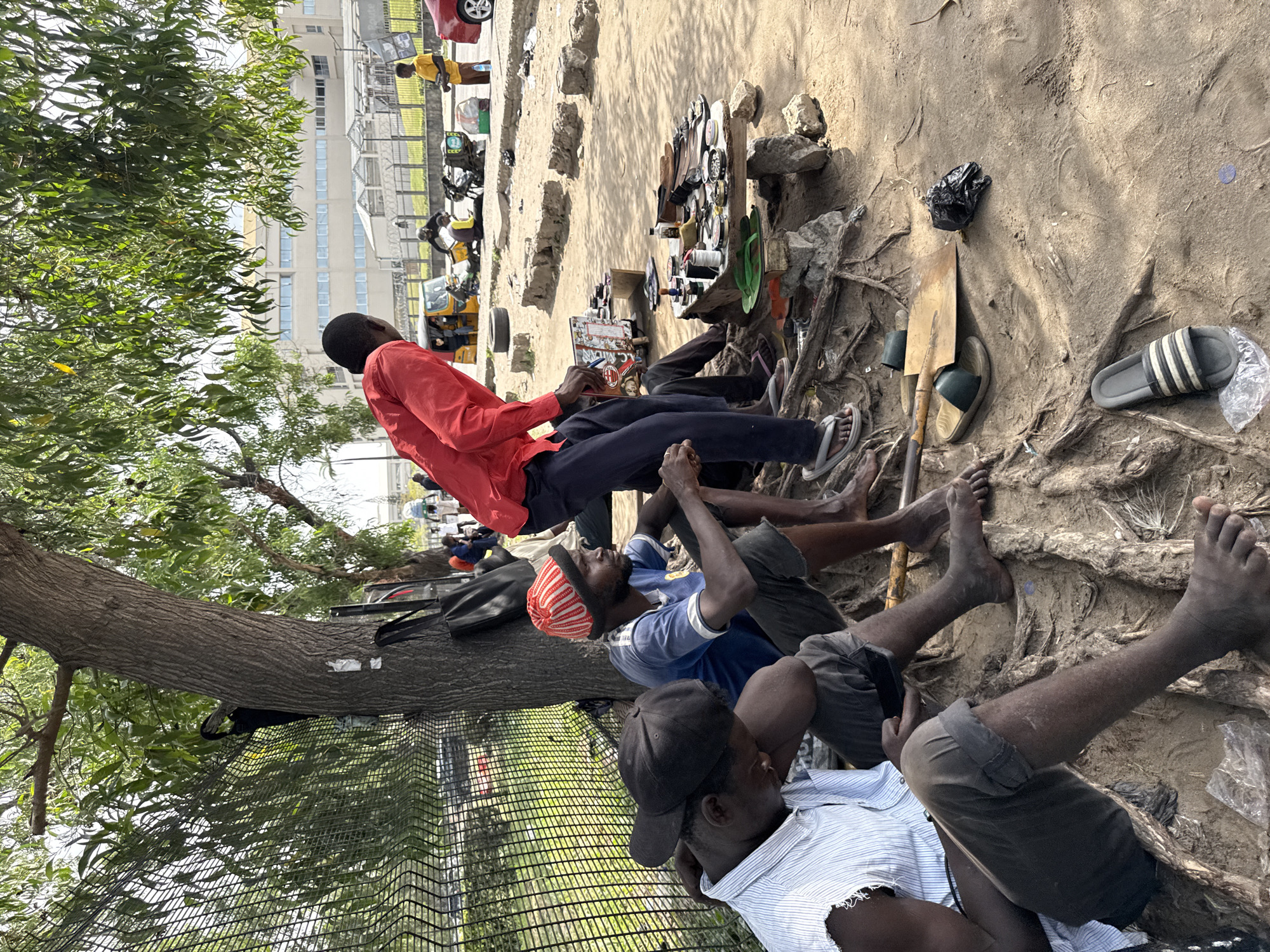 This image captures an outdoor scene in Igboefon, Nigeria, under bright daylight. Several men are gathered on sandy ground beneath large trees with dense green foliage.
In the foreground, a man in a red long-sleeved shirt and dark trousers is seated, looking down at a book or magazine with "C. MEAN" partially visible on its cover. He wears a flip-flop on his right foot. Near him, another man in a blue t-shirt and a red and white striped beanie is also seated. Further to the right, a man wearing a striped white and blue shirt and a black baseball cap is reclining on the ground, his bare feet showing. The men appear to be resting or waiting.
The ground is unpaved, featuring exposed tree roots and scattered dirt. Various items are displayed on the ground and small wooden platforms, indicating a street vending or repair setup. These items include shoes, sandals (one pair of black and white striped sandals is prominent on the ground), watches, and small accessories. A shovel and a black plastic bag are also visible on the ground.
In the background, behind a chain-link fence, several vehicles, including tricycles (keke Napeps) and a red car, are parked. A multi-story building with numerous windows is visible beyond the fence. Another individual in a yellow shirt stands near the vehicles. The scene depicts a casual, informal gathering, possibly a market area or roadside stop.
