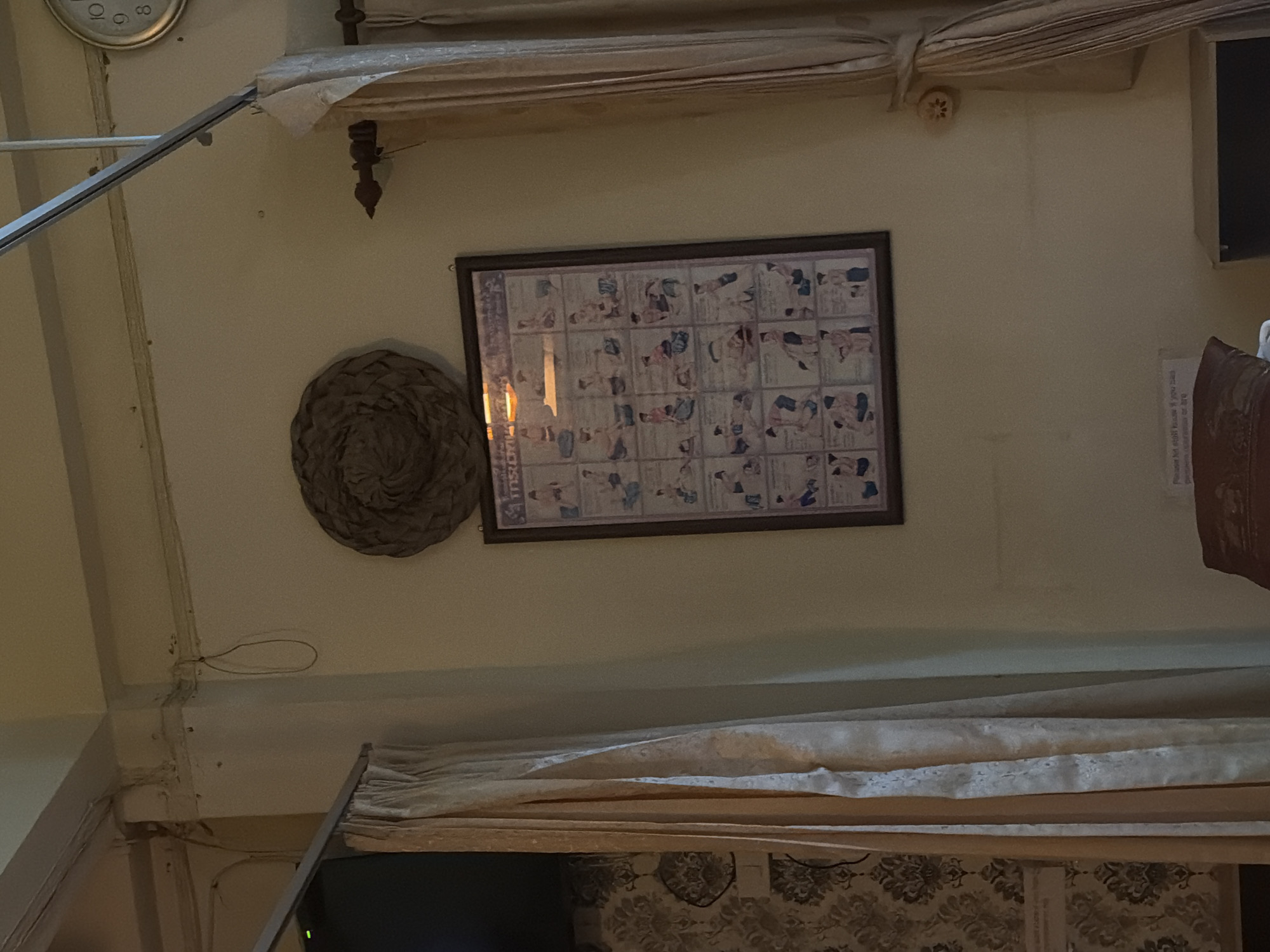 In Baan Tai, Thailand, a framed chart hangs on a cream-colored wall. The chart displays a grid of small, black and white illustrations depicting various massage techniques. Above the chart, a round, woven object is mounted. To the right of the chart, beige curtains hang from a curtain rod. A portion of a clock is visible in the upper right corner of the image. The wall exhibits some signs of wear, with visible dust and wiring. To the left, more beige curtains frame a dark rectangular object, possibly a television screen. The lower portion of the image shows a patterned wallpaper on the left, and a brown pillow resting on a surface on the bottom.