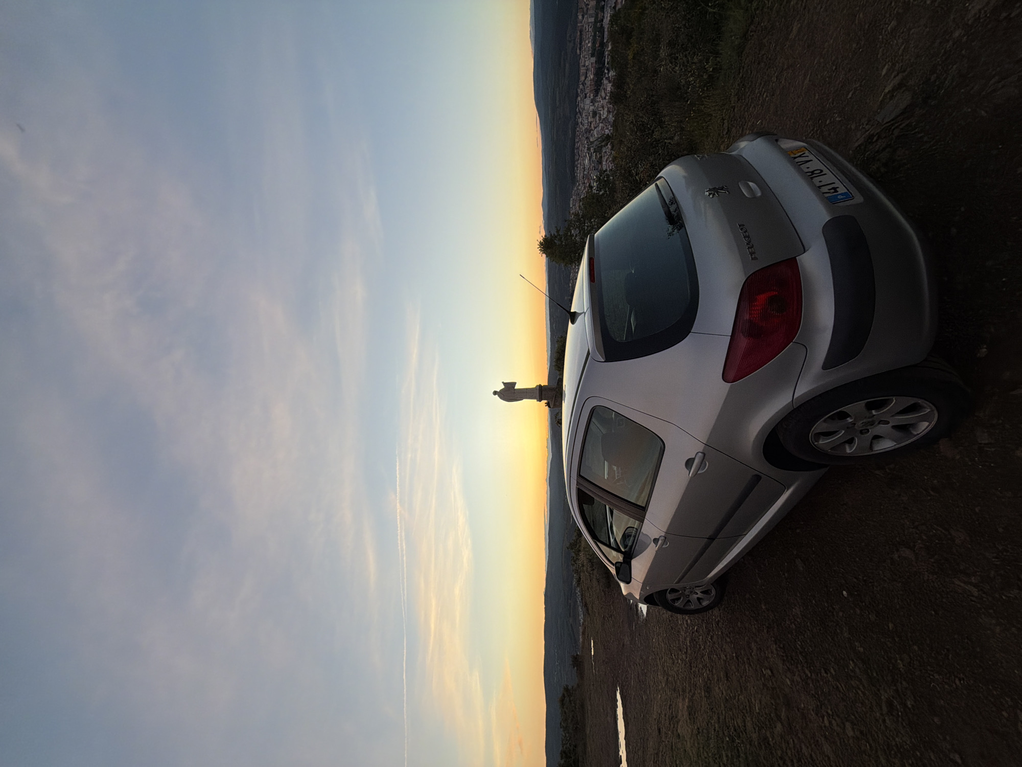 A silver Peugeot 207 is parked on a dirt road overlooking a city, at what appears to be the Sanctuary of Nossa Senhora do Castelo in Bragança, Portugal, as indicated by the statue of the Virgin Mary. The car's license plate is visible, showing "41-18-VX". The sun is setting, creating a golden glow in the sky. The terrain surrounding the car is earthy and uneven. The city lies in the valley below the viewpoint. The sky is a mix of blue and yellow hues.
