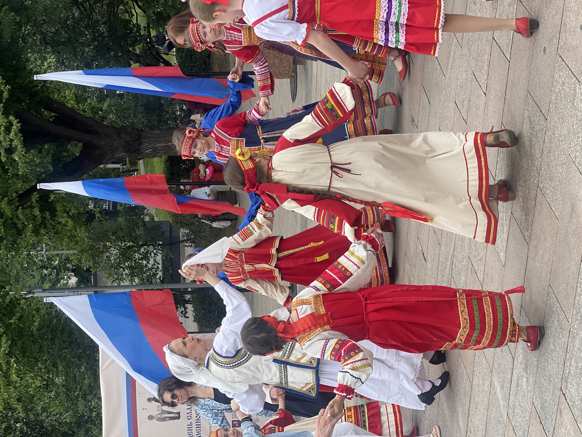A group of people are captured in motion, likely performing a traditional dance, while holding hands in a circle. They are dressed in colorful costumes. Russian flags surround the group. The setting appears to be outdoors on a paved square or plaza, with trees visible in the background. The location is Unknown, Unknown.