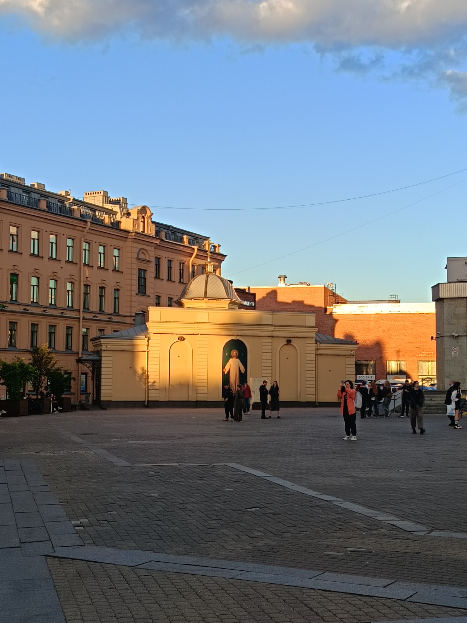 The image shows a street scene in Saint Petersburg, Russia, with a light pink building in the background and a small yellow structure in the foreground. A statue of a person, likely Jesus Christ, is visible within an arched alcove of the yellow structure. Several people are standing and walking in the paved plaza, with some appearing to take photographs. The sky is clear blue with some clouds, and the sun is casting long shadows, suggesting it is late afternoon or early evening. The plaza is paved with cobblestones and interlinked grey stone tiles. The overall impression is of a public square with a religious or historical monument.