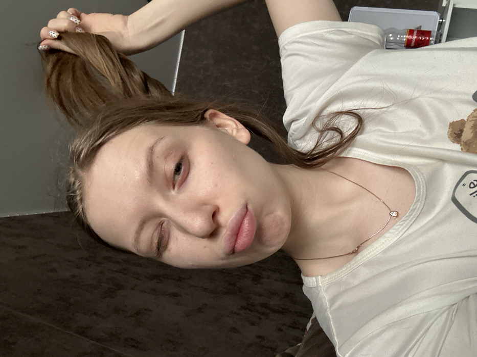 A portrait-oriented image depicts a young female subject in what appears to be an indoor setting in the city of Chaikovsky, Russia. The subject, with light skin and brown hair, is looking directly at the camera. Her left eye is closed in a wink, while her right eye is partially open. Her lips are pursed forward in a "duck face" expression. Her right hand is raised, holding a substantial portion of her brown hair upwards. The fingernails on her right hand feature a white polish with small black dots. She is wearing a white short-sleeved t-shirt, revealing a partial graphic design on the lower right, possibly depicting a cartoon bear. A thin, gold-colored necklace with a small, clear, teardrop-shaped pendant is visible around her neck. The background consists predominantly of a dark brown, textured wall. To the far right, a portion of a light-colored surface, possibly a shelf or appliance, is visible, upon which rests a clear plastic bottle with red labeling.