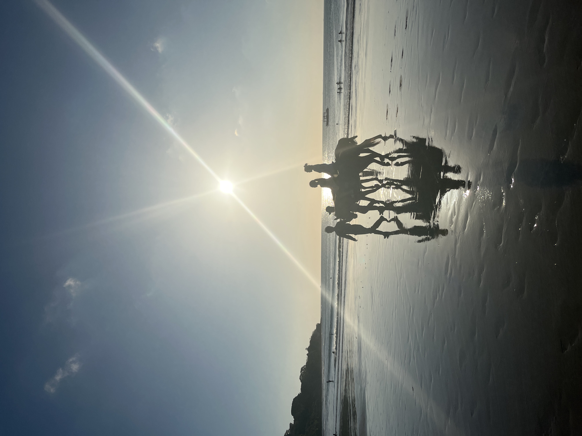 An extreme wide shot captures a beach scene at sunset in Kuta Selatan, Indonesia, with the sun creating a prominent lens flare across the sky and reflecting on the wet sand. In the foreground, silhouetted figures are visible. A group of three individuals are riding on horseback, with two figures riding one horse and another person walking alongside. To the right of the horse riders, two additional silhouetted figures walk together. In the background, the ocean is calm with small waves breaking on the shore. Several people are visible in the water, swimming or wading. A distant boat is on the horizon. On the left side of the image, a headland with vegetation juts out into the sea. The wet sand in the foreground reflects the sky and the figures, creating a mirror-like effect.